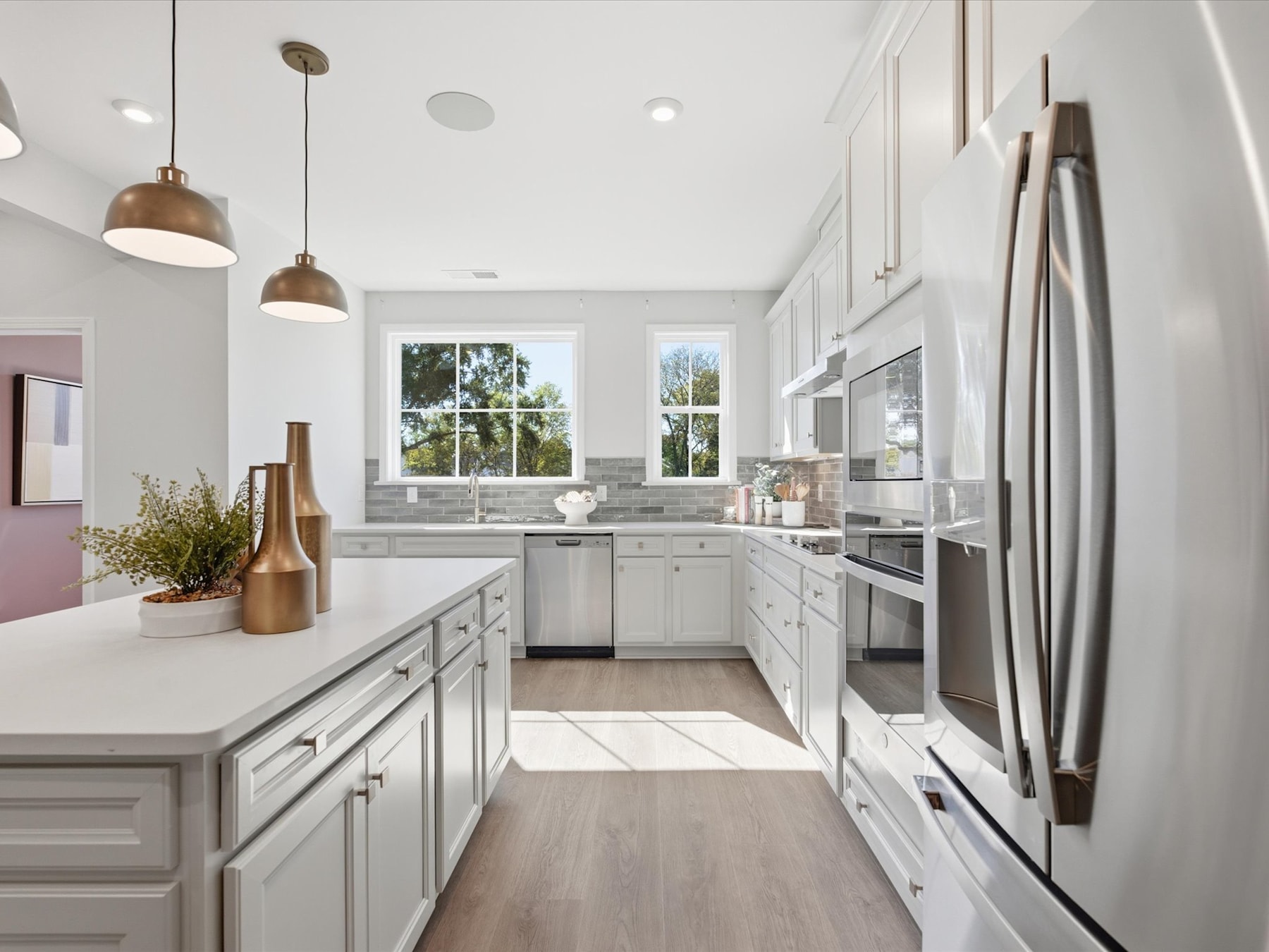 A modern, bright, and spacious kitchen with white cabinets, stainless steel appliances, and pendant lighting fixtures, set against a backdrop of large windows overlooking an outdoor scene.