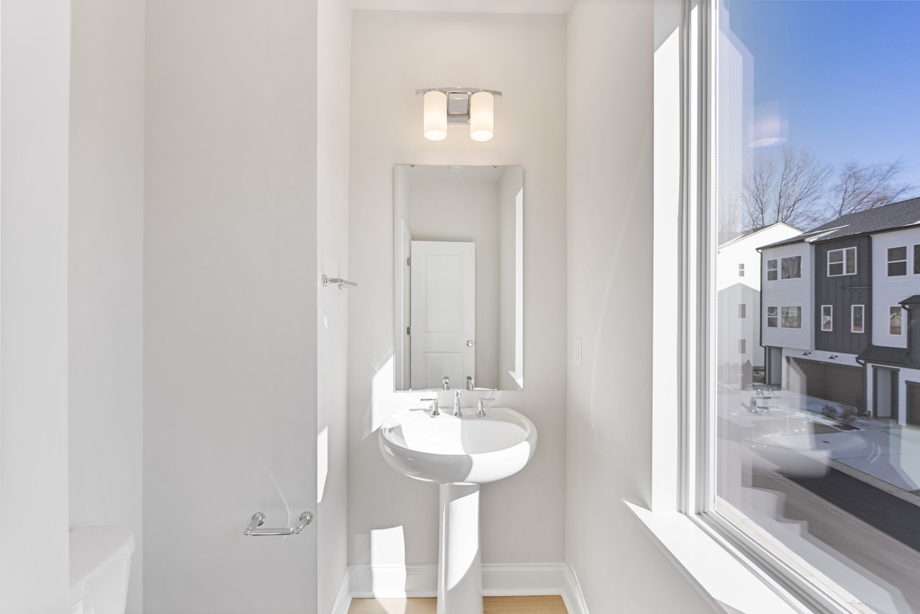 A bright and minimalist bathroom with a white sink and mirror, surrounded by a window overlooking a residential neighborhood outside.