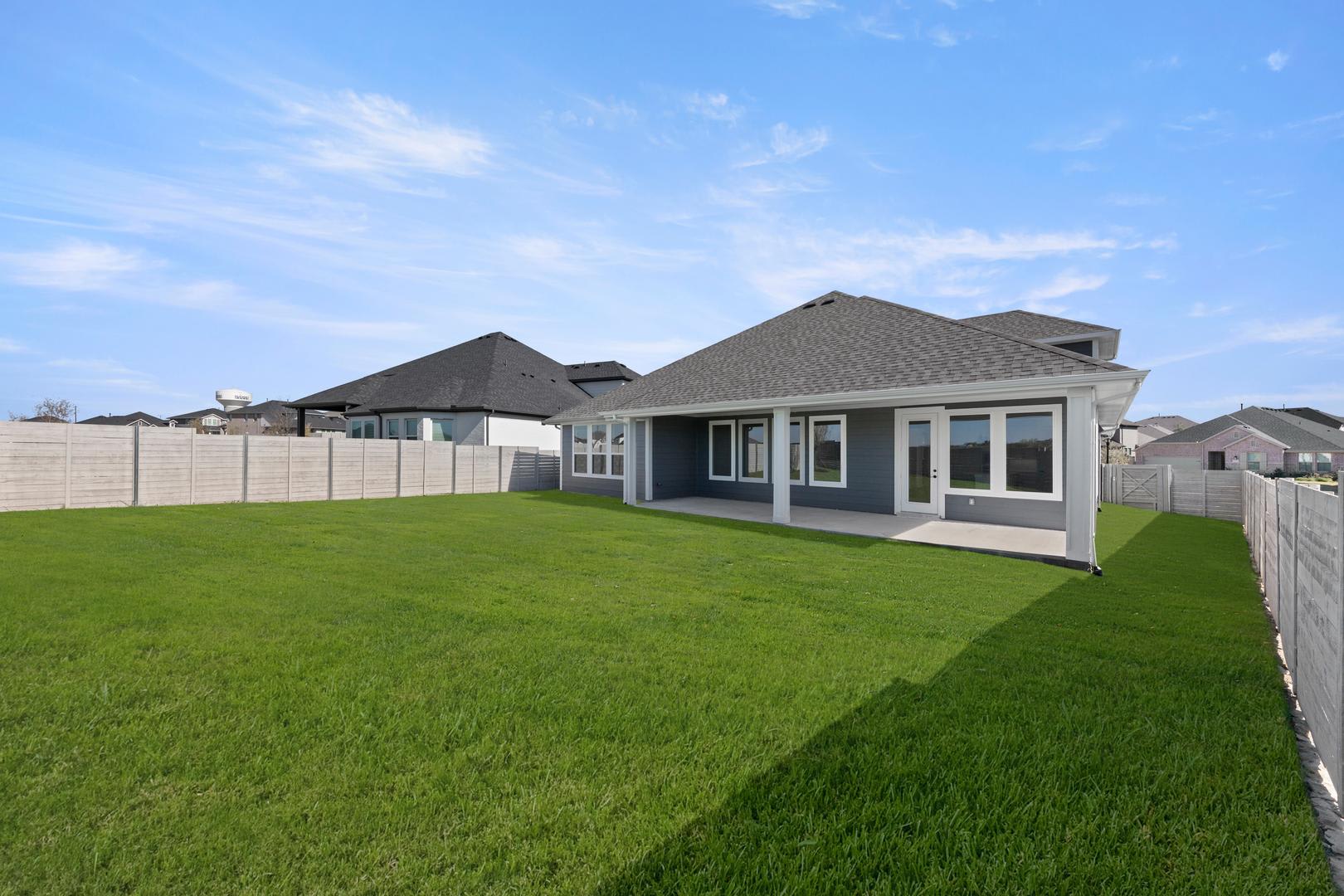 A large, modern house with a gray roof and siding sits on a well-manicured lawn, surrounded by a wooden fence, against a clear blue sky with wispy clouds.