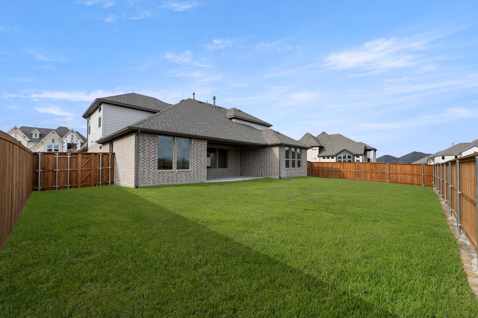 A well-manicured lawn stretches out in the foreground, leading to a two-story gray house with a slanted roof and a wooden fence surrounding the property in the background.
