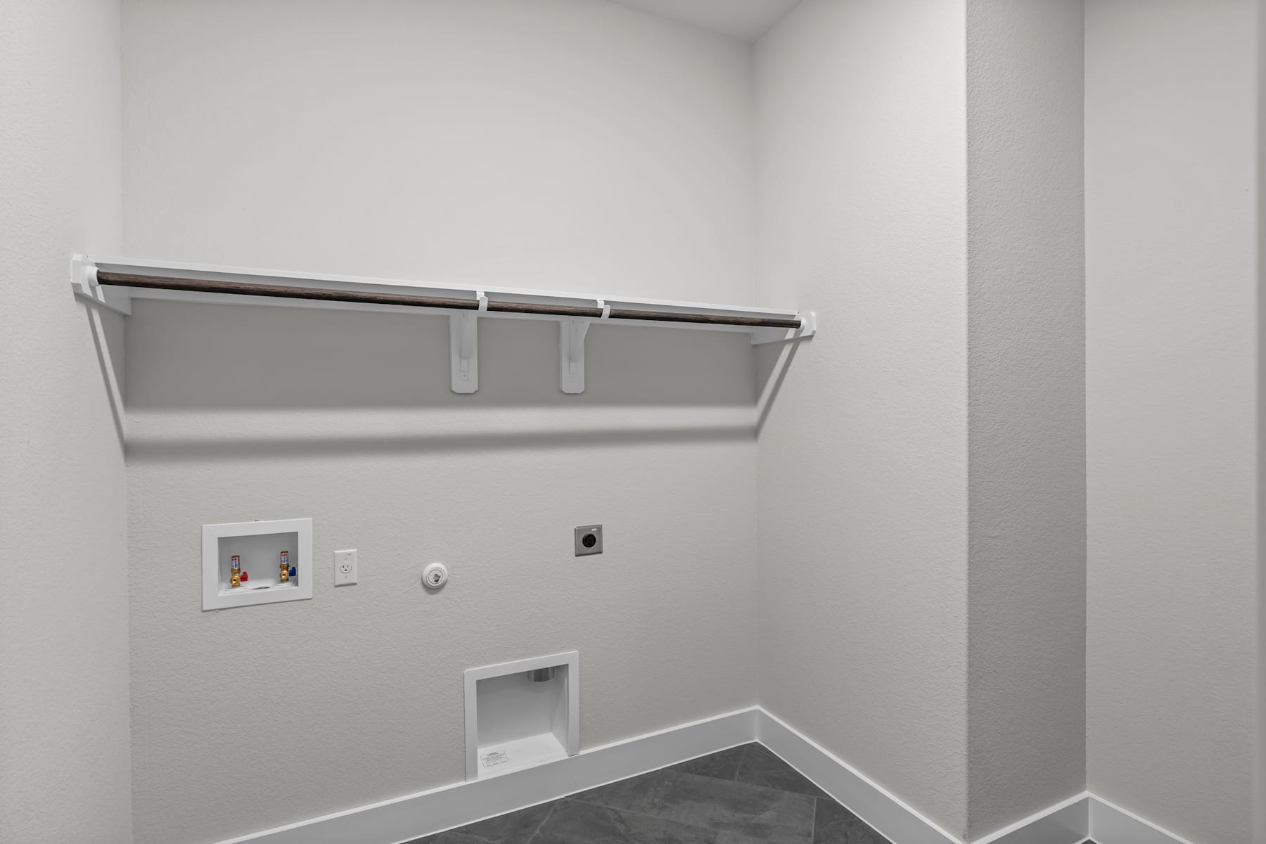 A minimalist, white-walled room with a simple clothing rod and electrical outlets visible.