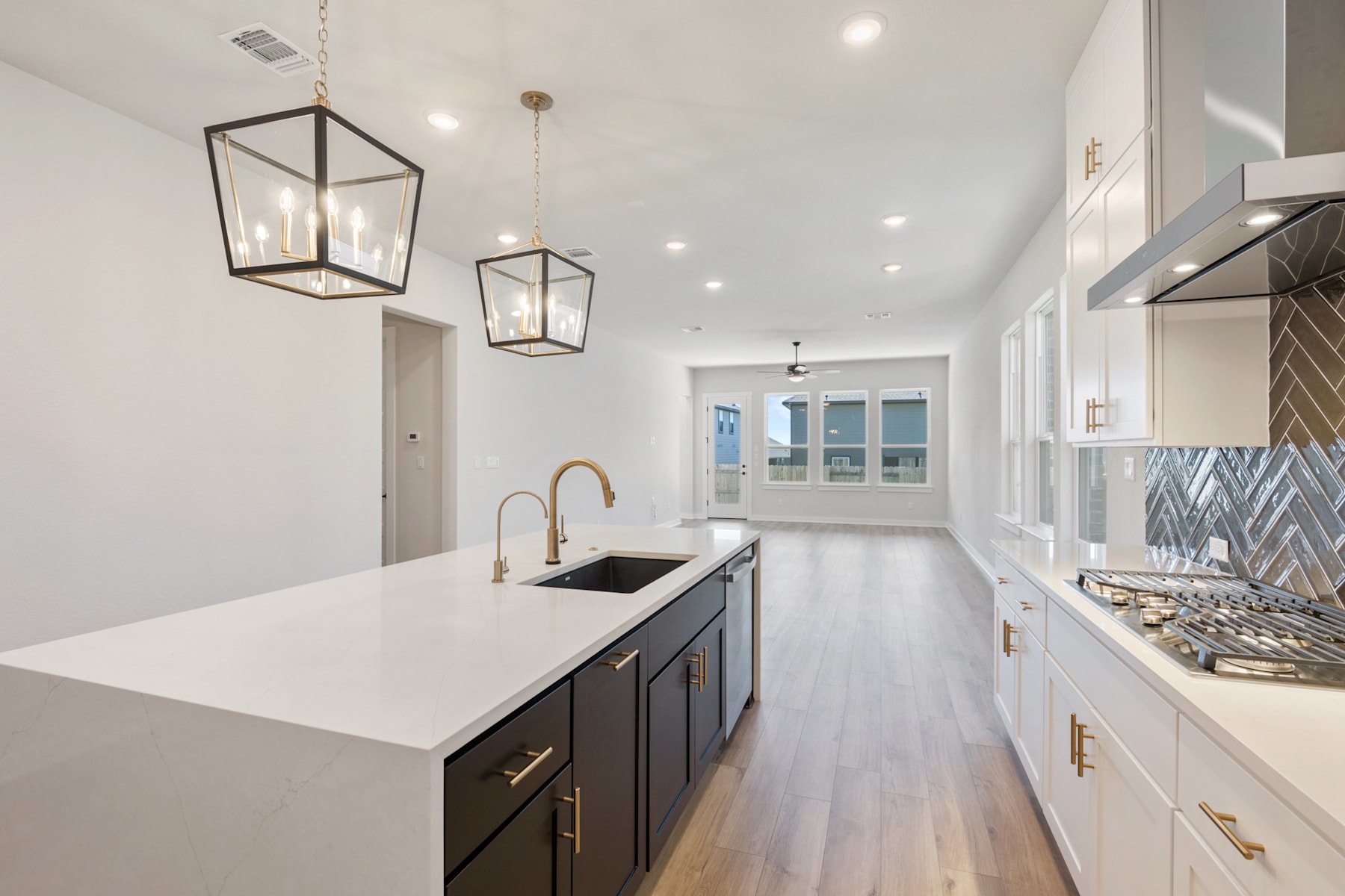 A modern, well-lit kitchen with sleek white countertops, dark cabinets, and pendant lighting fixtures, leading into a bright and airy hallway.