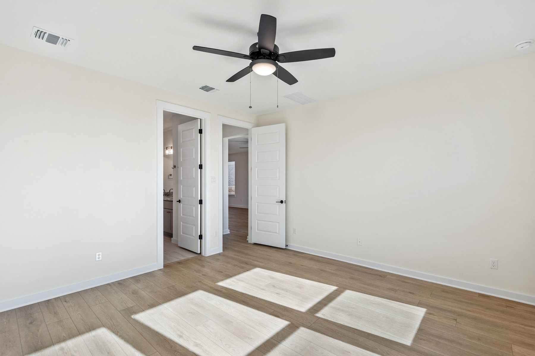 A spacious room with a ceiling fan, white walls, and a hardwood floor with a geometric pattern of light and shadow.
