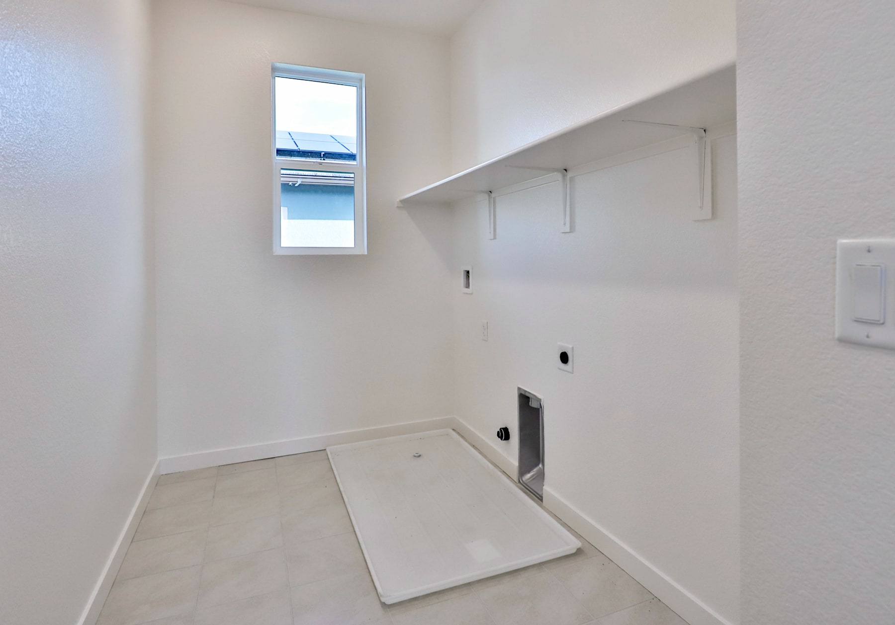 A bright, minimalist laundry room with a white tiled floor, a small window, and a recessed wall space for a washer and dryer.