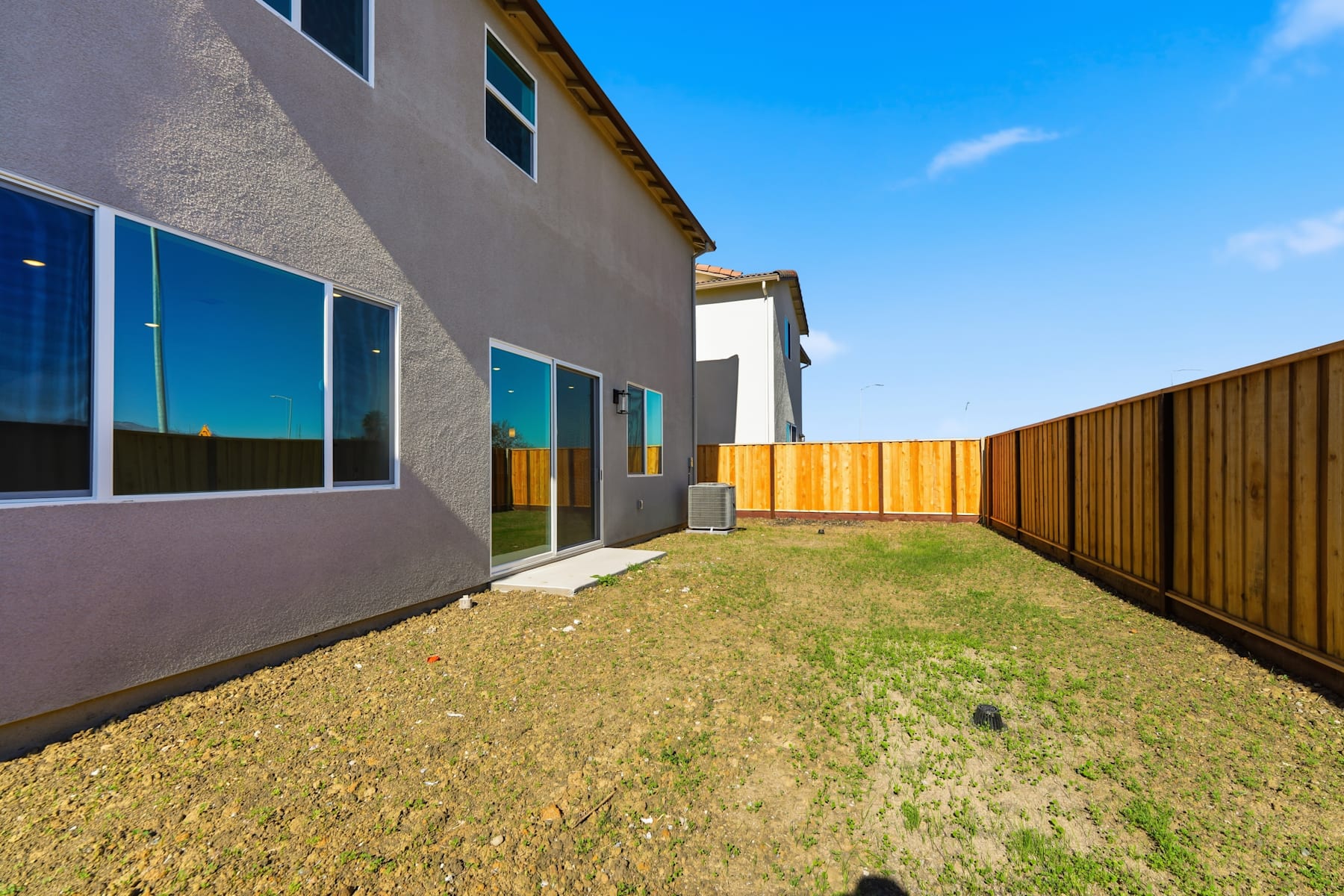 A modern, two-story residential building with a wooden fence and a grassy yard in the foreground, set against a clear blue sky.
