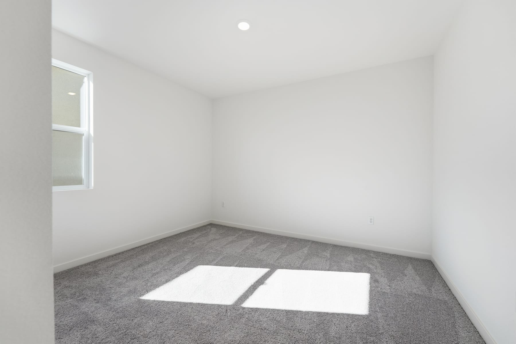 An empty room with white walls, a gray carpeted floor, and a window letting in natural light, creating a bright and minimalist atmosphere.