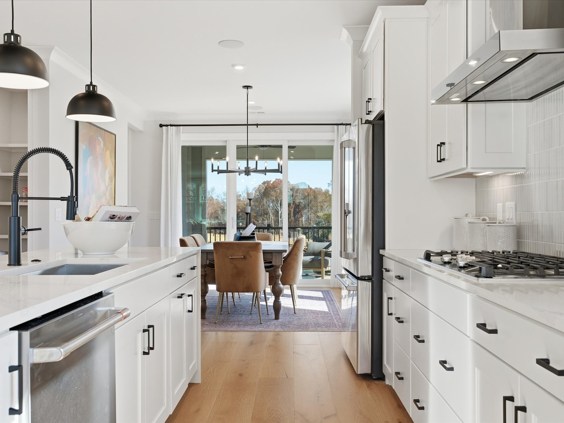 A modern, bright kitchen with white cabinets, stainless steel appliances, and a dining area visible in the background through an open doorway.