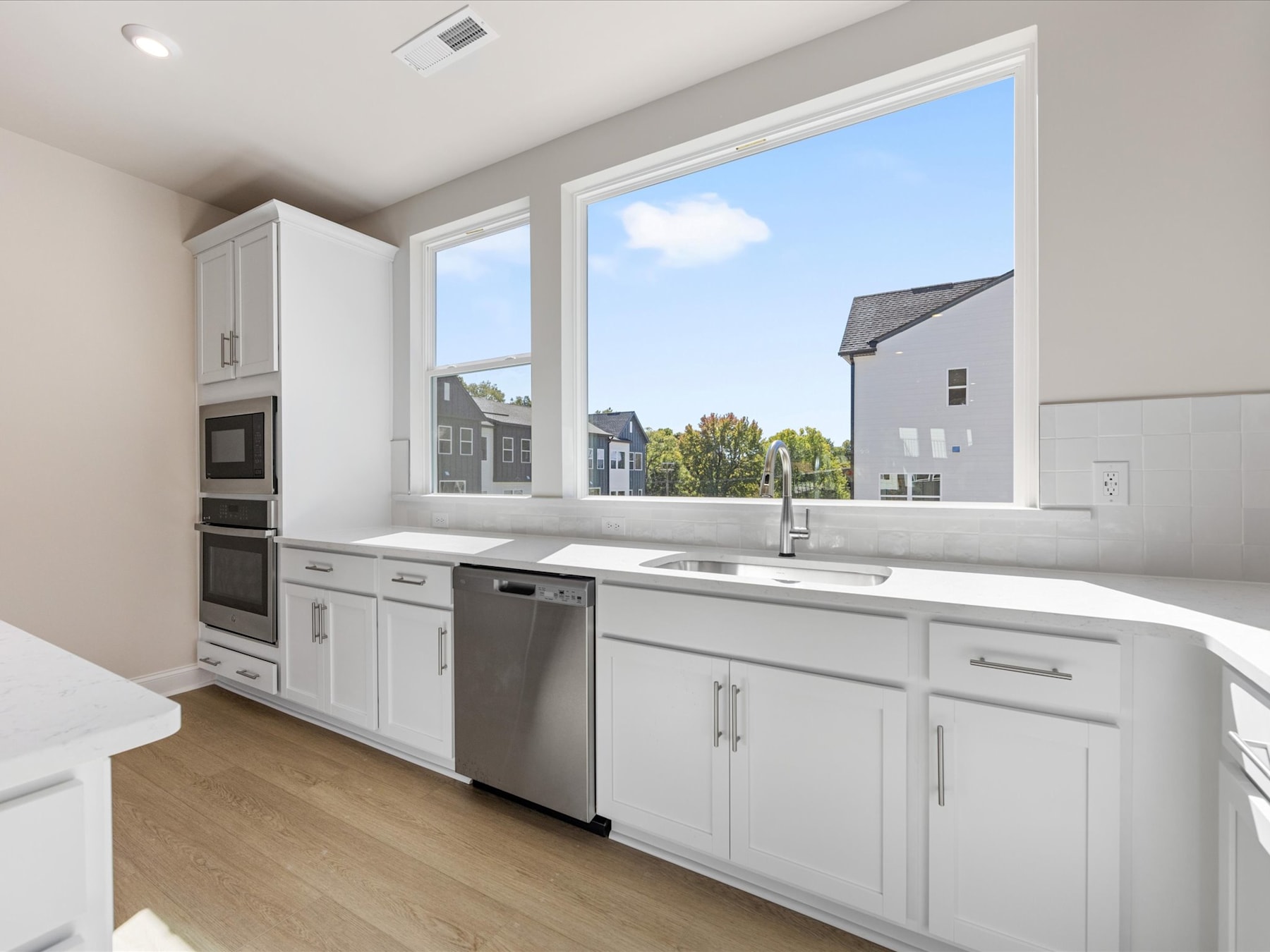 A modern, well-lit kitchen with white cabinets, stainless steel appliances, and a view of residential buildings in the background through large windows.