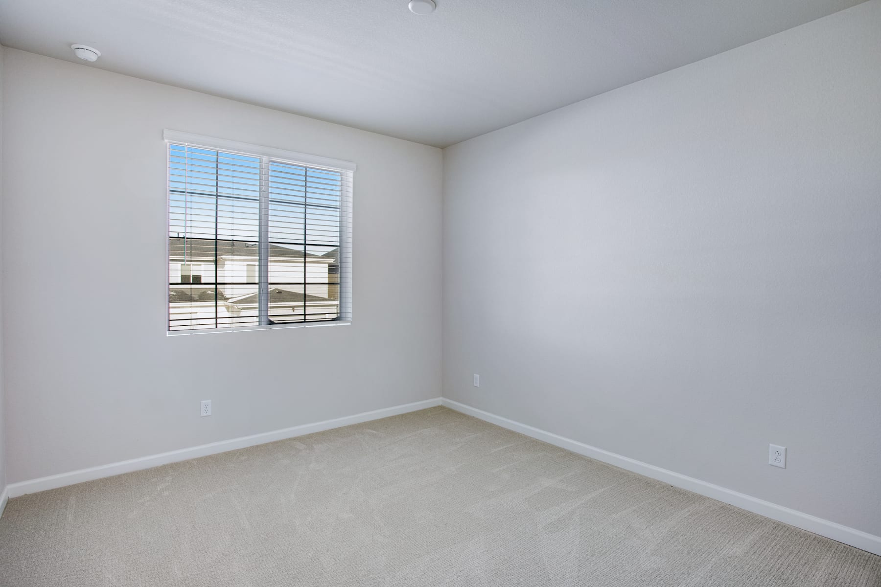 A bright, empty room with a large window overlooking a residential area, featuring white walls and a carpeted floor.