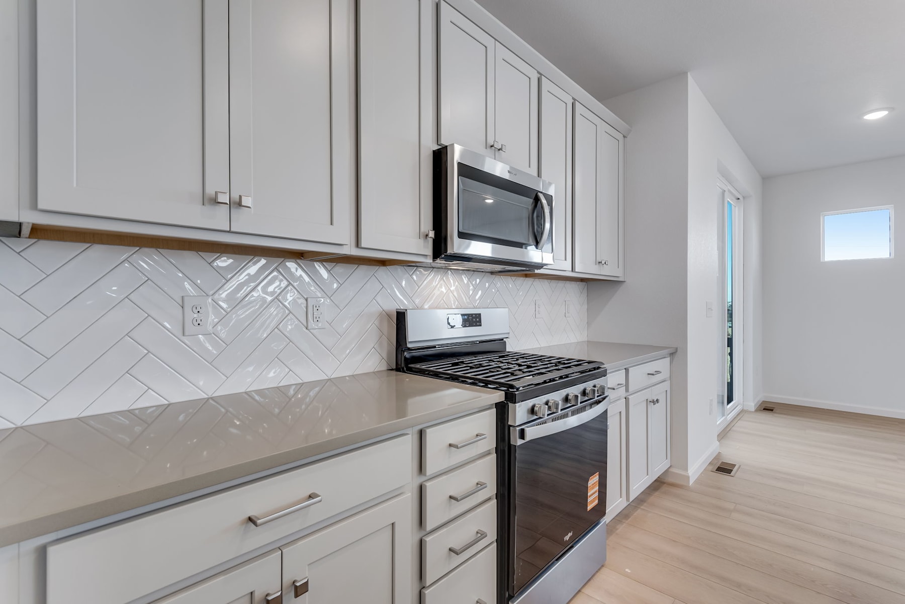 A modern, well-equipped kitchen with gray cabinets, a stainless steel oven, and a herringbone-patterned backsplash, set against a light-colored hardwood floor.