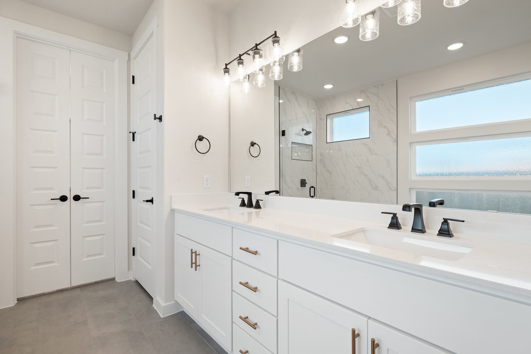 A modern, well-lit bathroom with white cabinets, a marble countertop, and a large mirror adorned with track lighting.