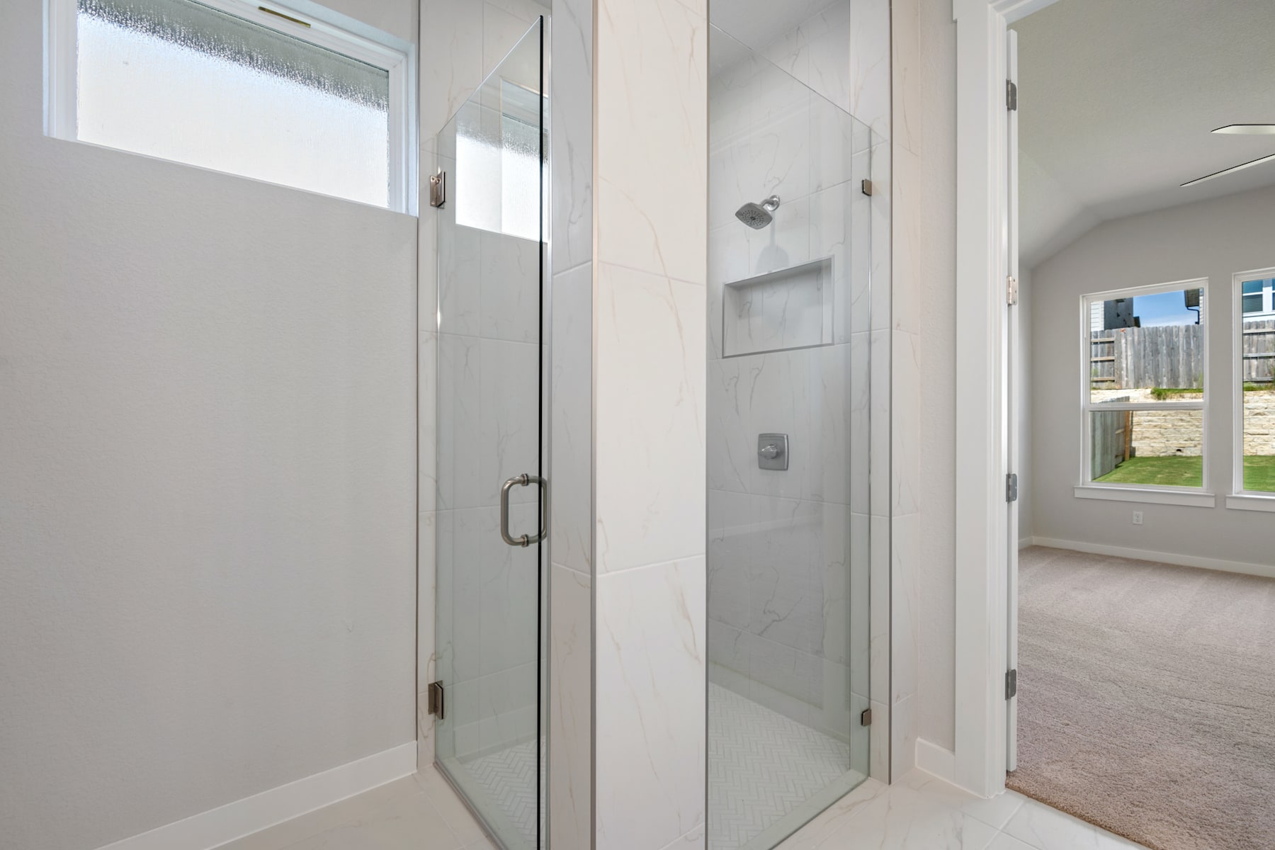 A modern, minimalist bathroom with a glass shower enclosure, white walls, and a window providing natural light.