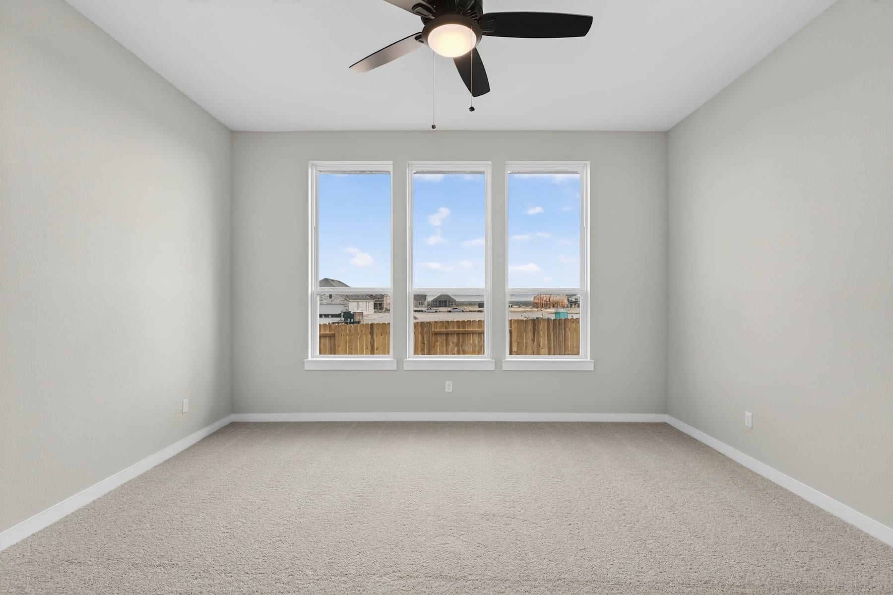 A sparsely furnished room with a ceiling fan, large windows overlooking a backyard, and a plain, carpeted floor.