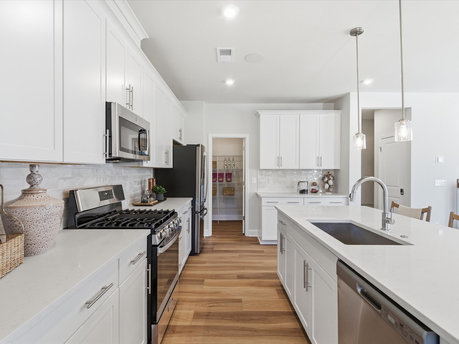 A modern, bright kitchen with white cabinets, stainless steel appliances, and a wooden floor extends into a hallway in the background.