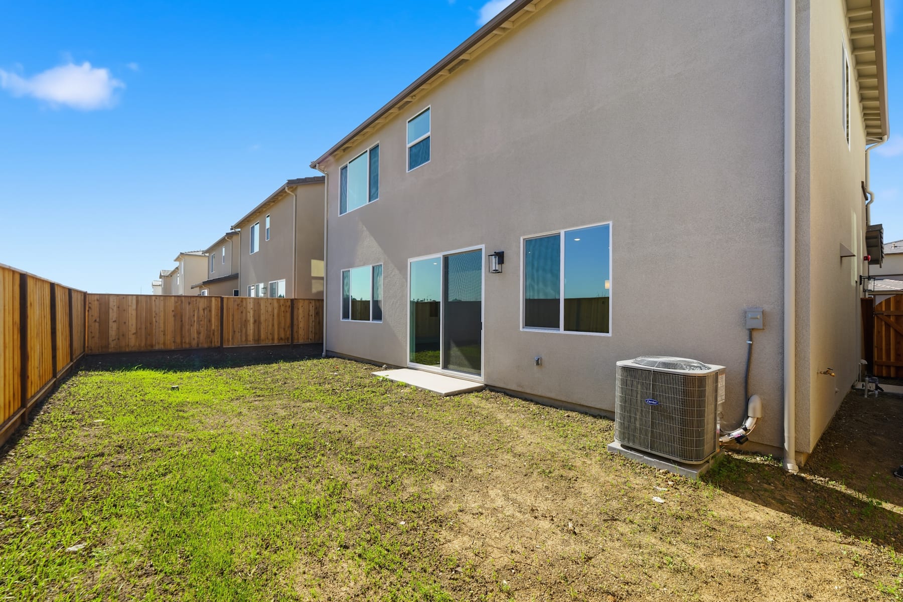 A newly constructed townhouse with a small grassy yard and a fenced backyard, set against a clear blue sky.