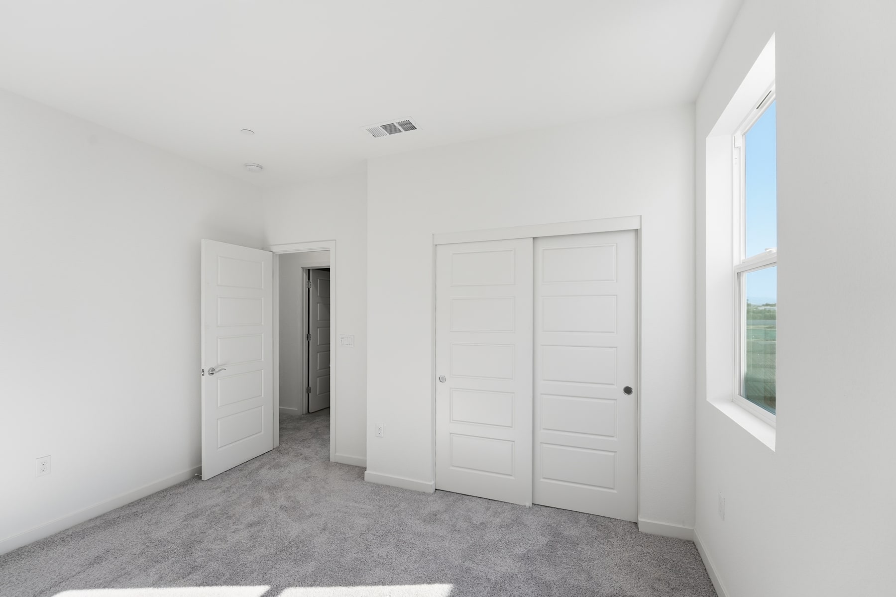 A spacious, minimalist bedroom with white walls, gray carpeting, and a large window providing natural light. The room features two white doors, likely leading to a closet and hallway.