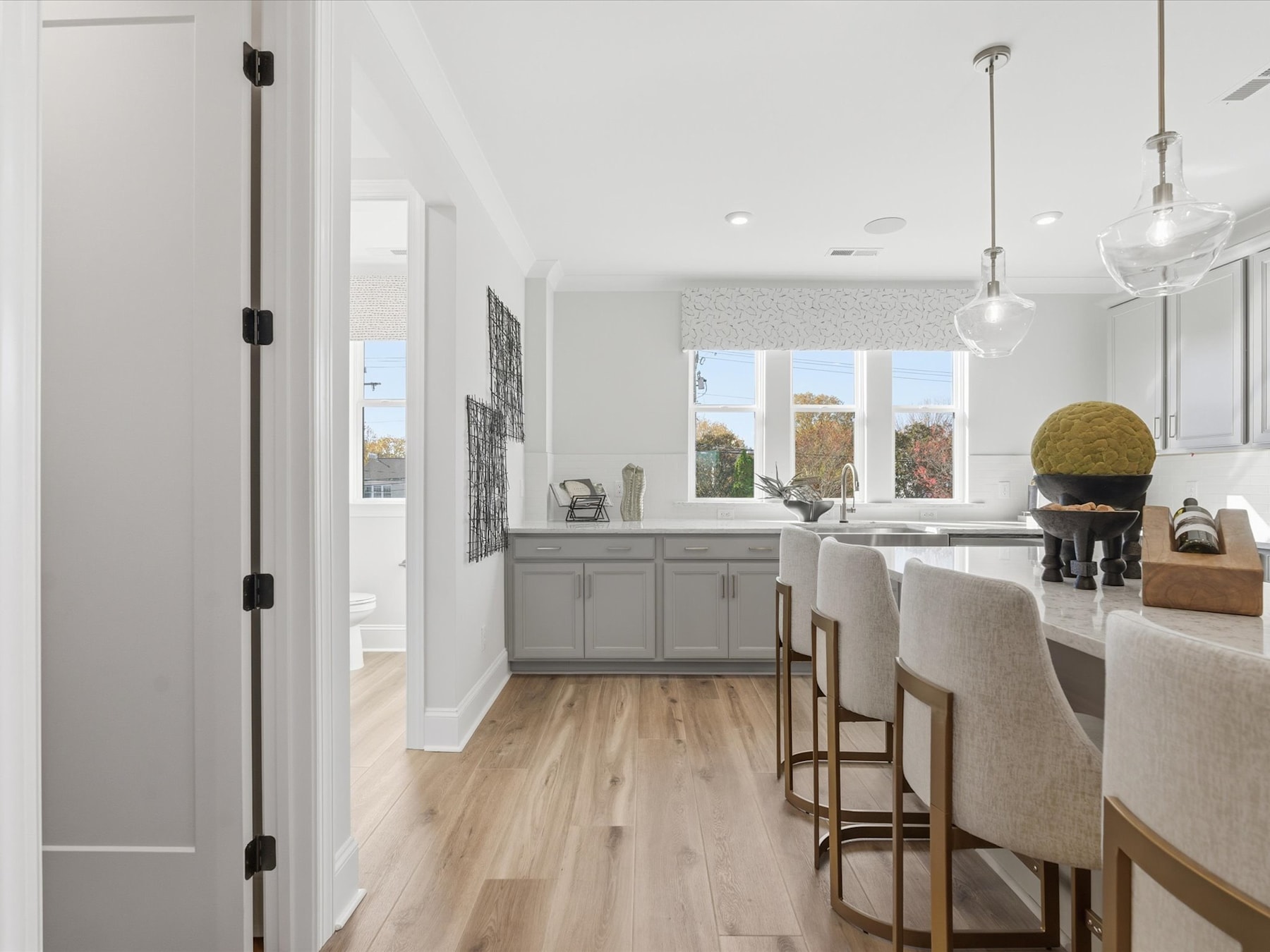 A modern, open-concept kitchen with white cabinets, wooden floors, and pendant lighting, overlooking a scenic outdoor view through large windows.