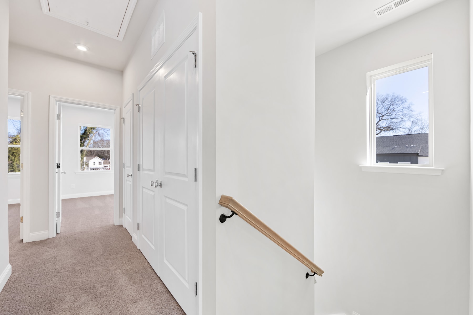 A bright, spacious hallway with white walls, a wooden handrail, and a window overlooking the exterior of the building.
