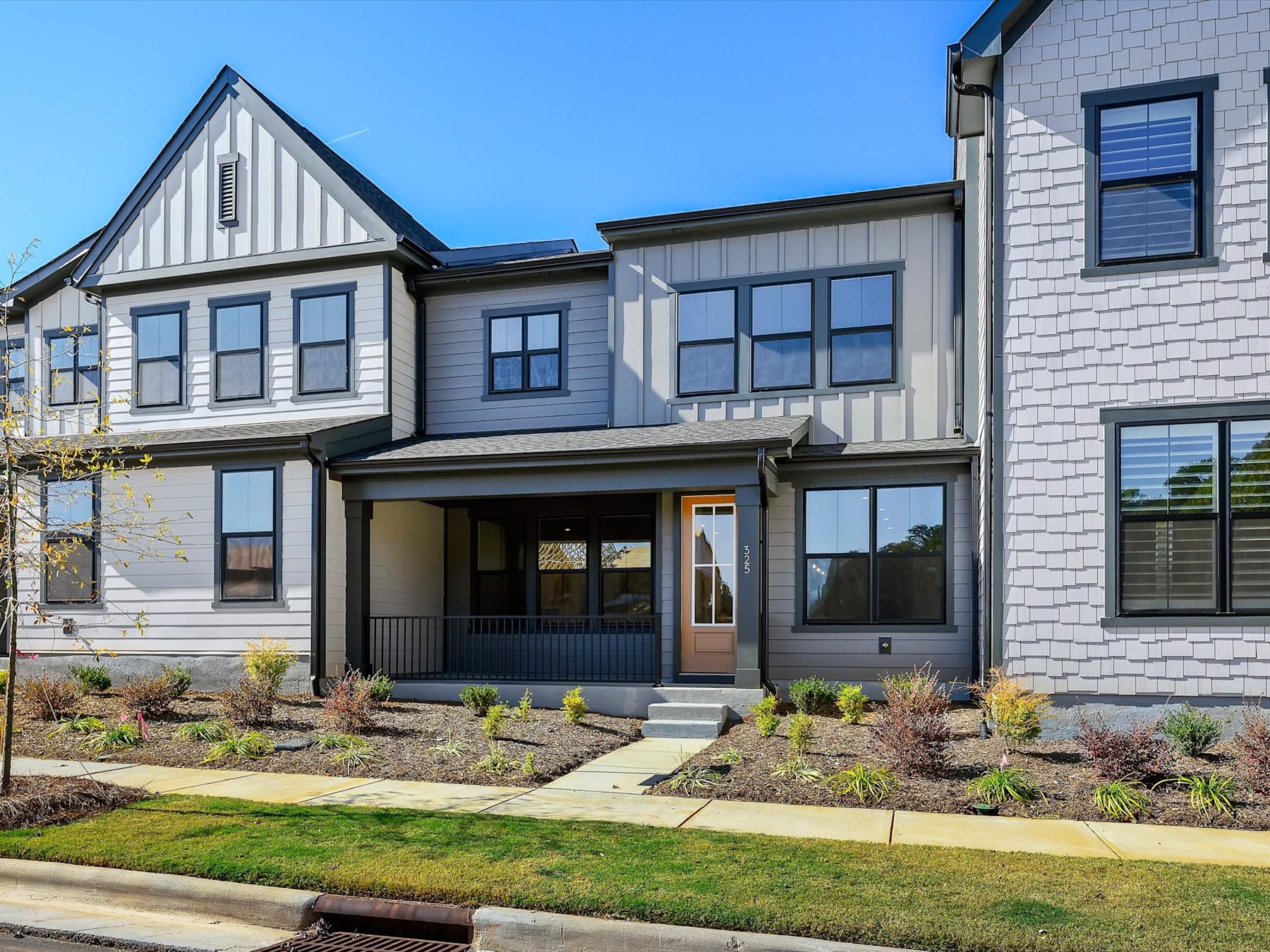 A modern, multi-story townhouse with a well-landscaped front yard, featuring a mix of siding materials, large windows, and a covered entryway.