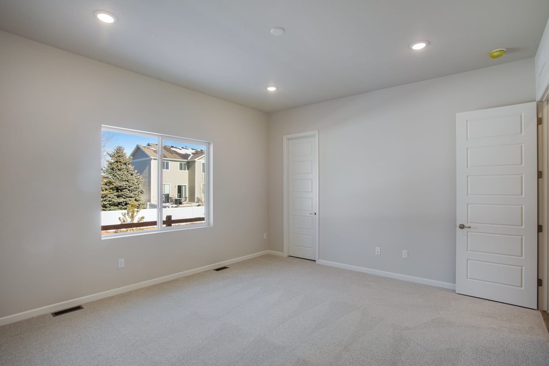 A spacious, well-lit room with a large window overlooking a snowy outdoor scene, featuring a white door and neutral-toned walls and flooring.