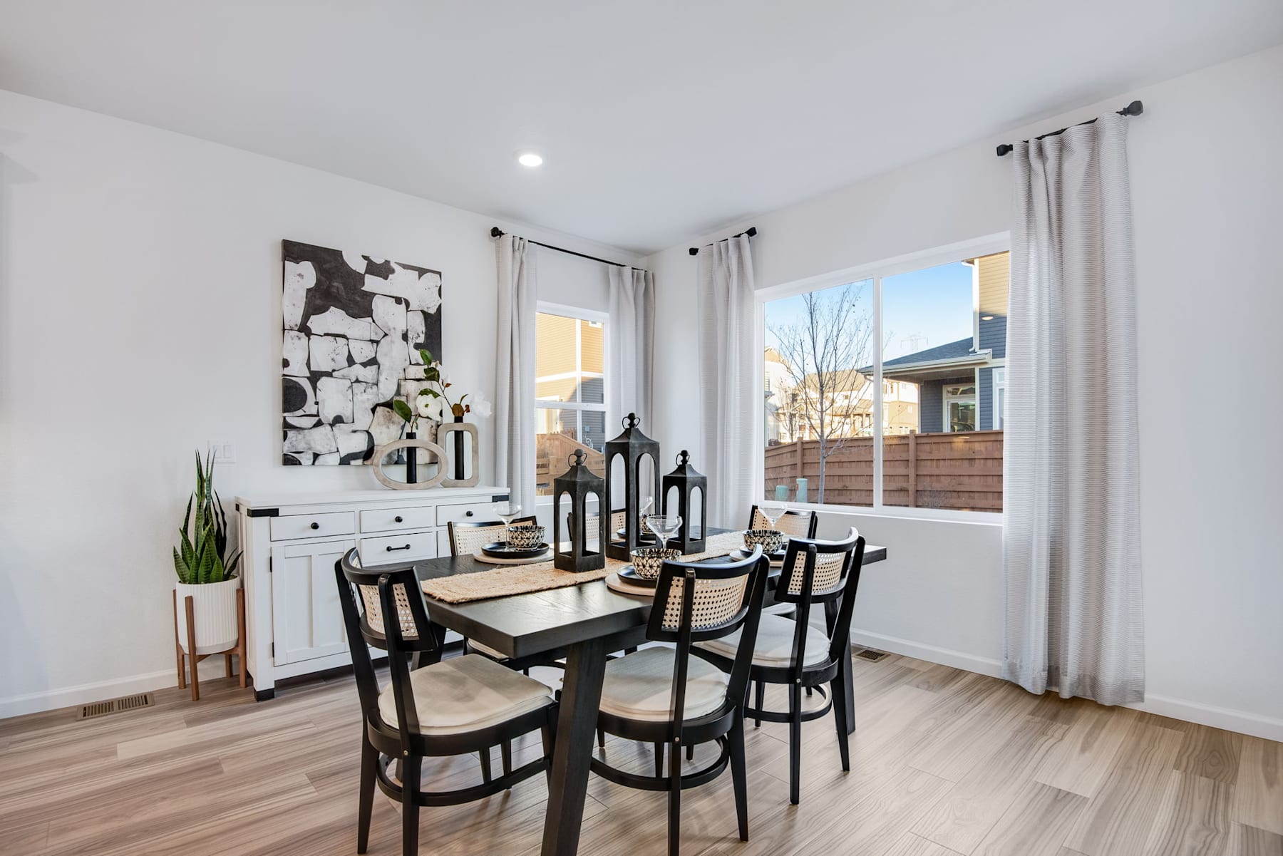 A modern and minimalist dining room with a wooden table, black chairs, and a large abstract artwork on the wall, surrounded by large windows that let in natural light.