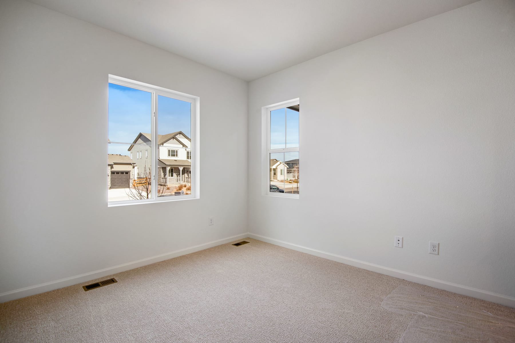An empty room with white walls, a carpeted floor, and large windows overlooking a residential neighborhood in the background.