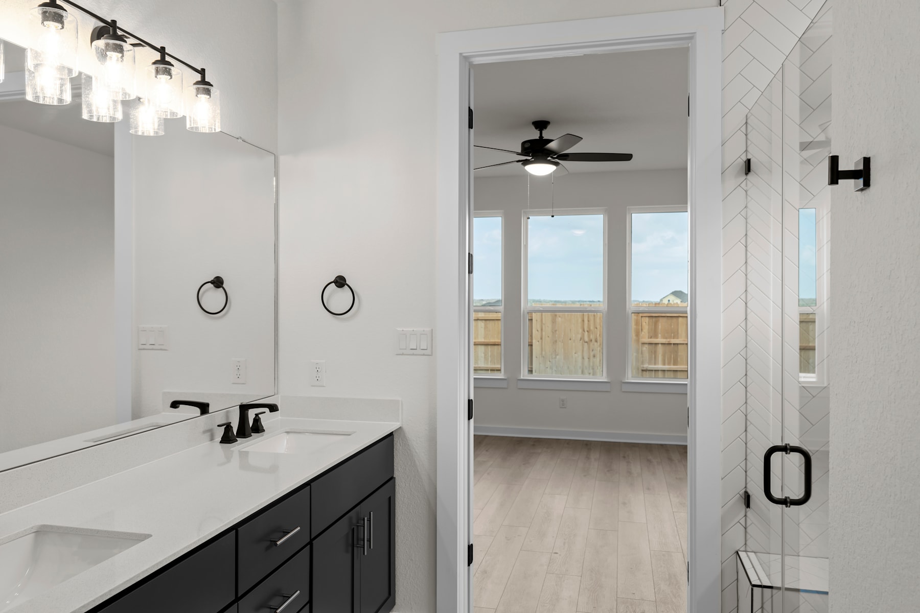 A modern and well-lit bathroom with a double vanity, black cabinets, and a ceiling fan visible in the adjacent room.