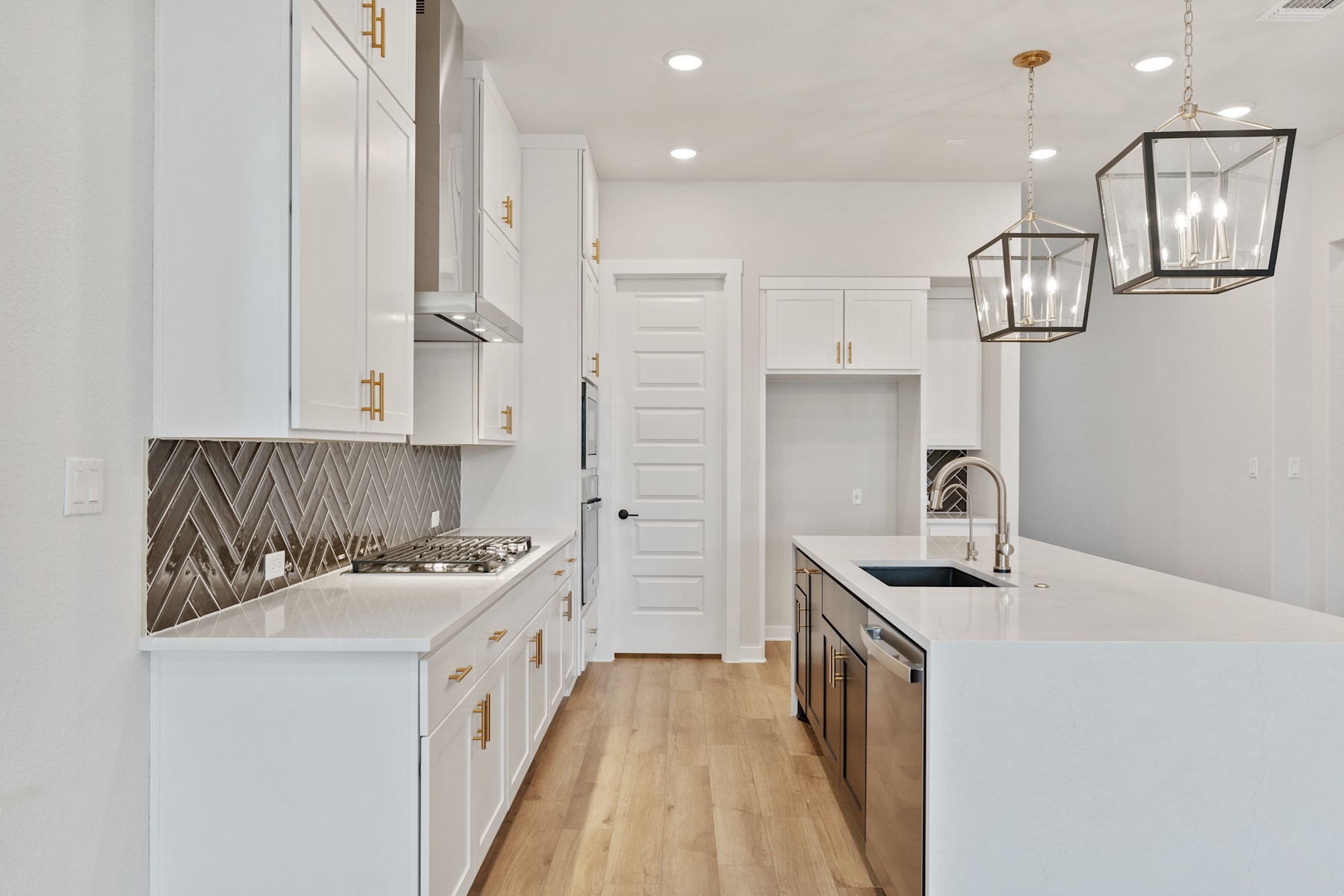 A modern, well-lit kitchen with white cabinets, a chevron-patterned backsplash, and sleek stainless steel appliances, set against a hardwood floor and accented by stylish pendant lighting.