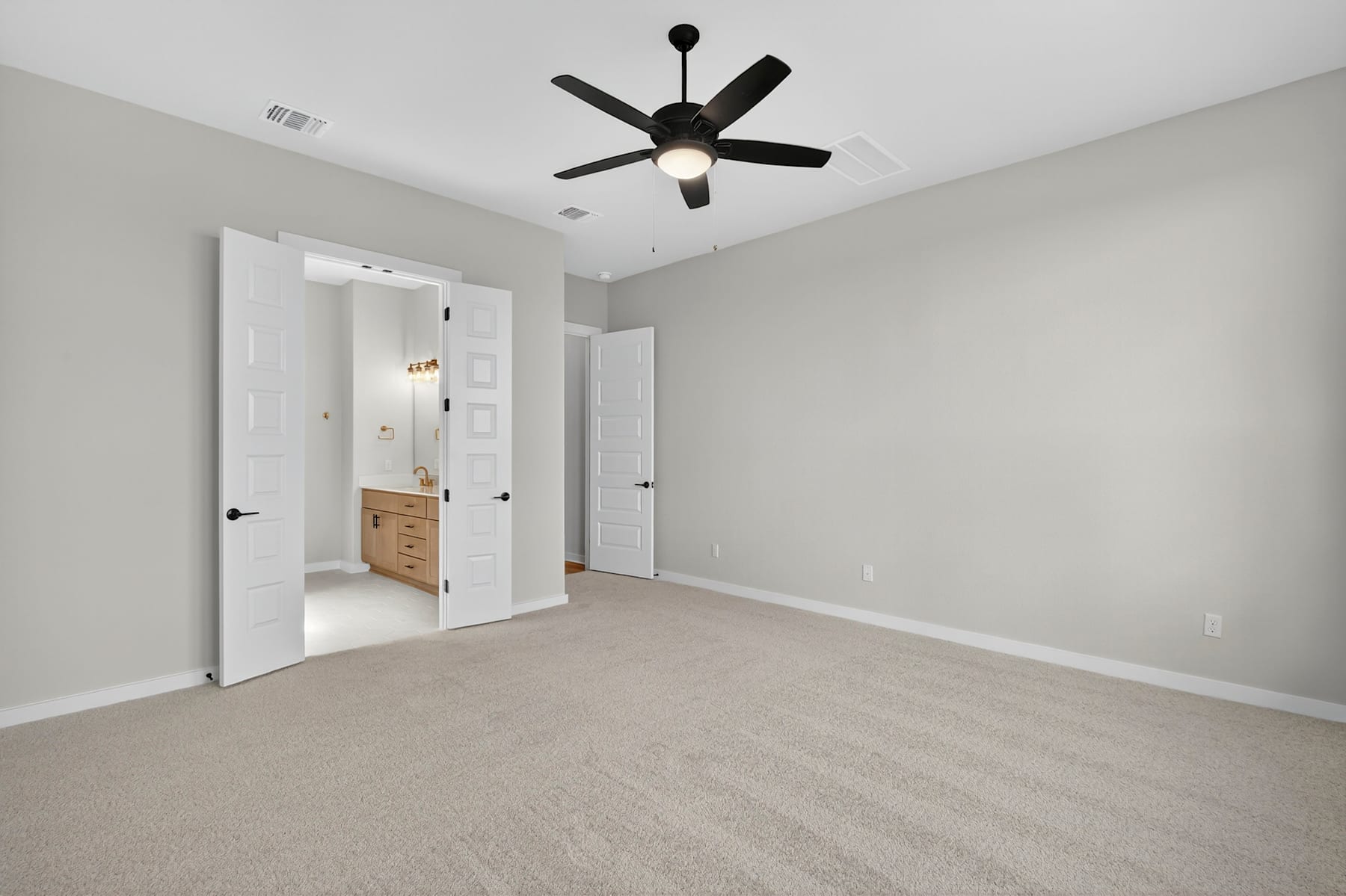 A spacious, minimalist bedroom with a ceiling fan, white walls, and beige carpeting, featuring an open doorway leading to another room.