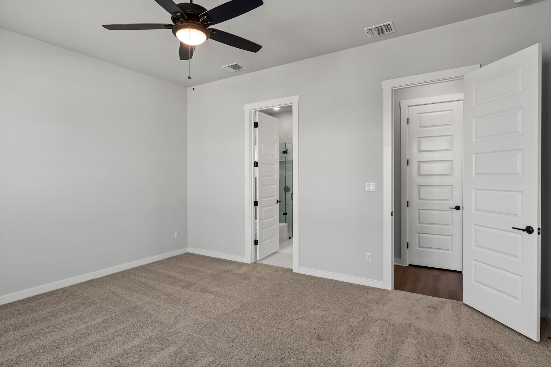 A spacious, well-lit room with a ceiling fan, white walls, and a carpeted floor. The room features a doorway and a mirror, creating a sense of depth and openness.