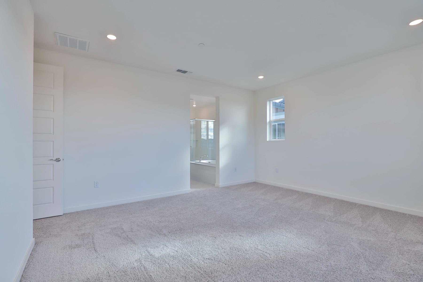 A spacious, well-lit room with white walls, a carpeted floor, and a doorway leading to another area.