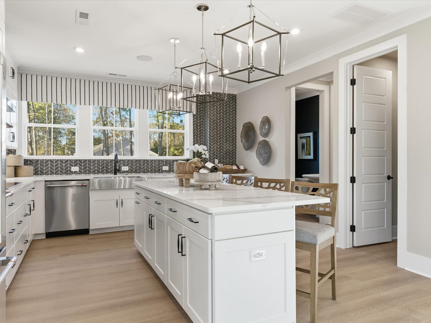 A modern, well-lit kitchen with white cabinets, a large island, and pendant lighting fixtures, set against a backdrop of large windows overlooking a scenic outdoor area.