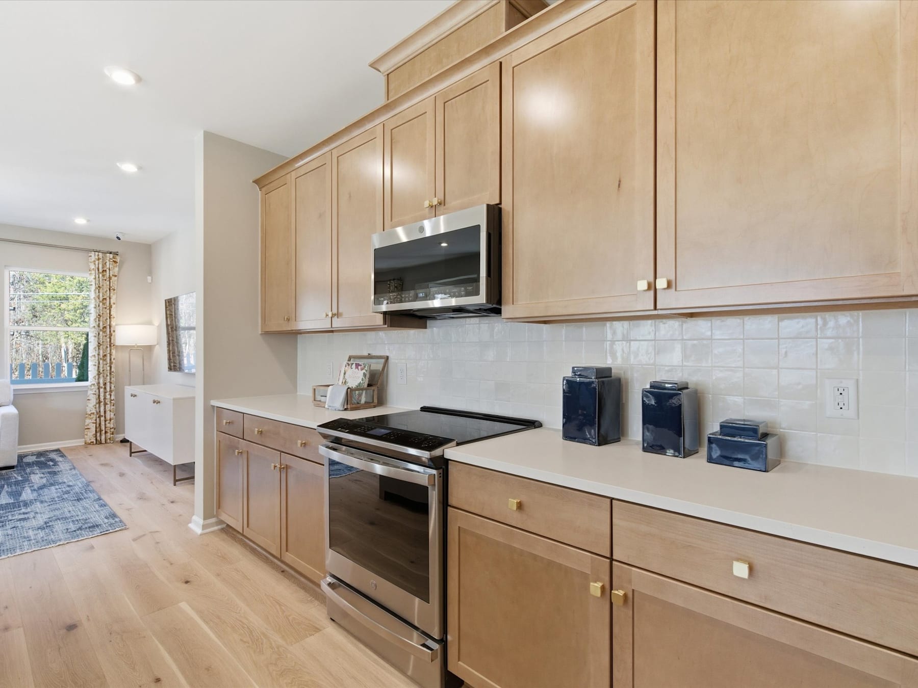 A modern kitchen with light wood cabinets, stainless steel appliances, and a tiled backsplash, set against a hardwood floor and a window overlooking a scenic outdoor view.