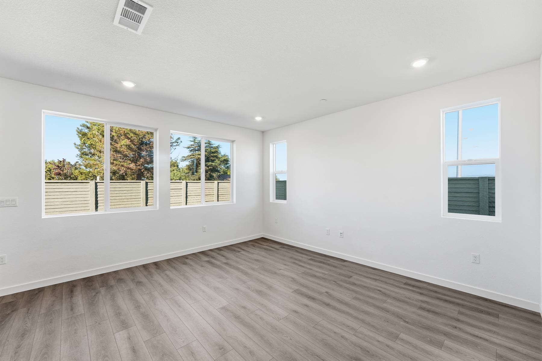 A bright, spacious room with large windows overlooking a wooded area, featuring a hardwood floor and recessed lighting.