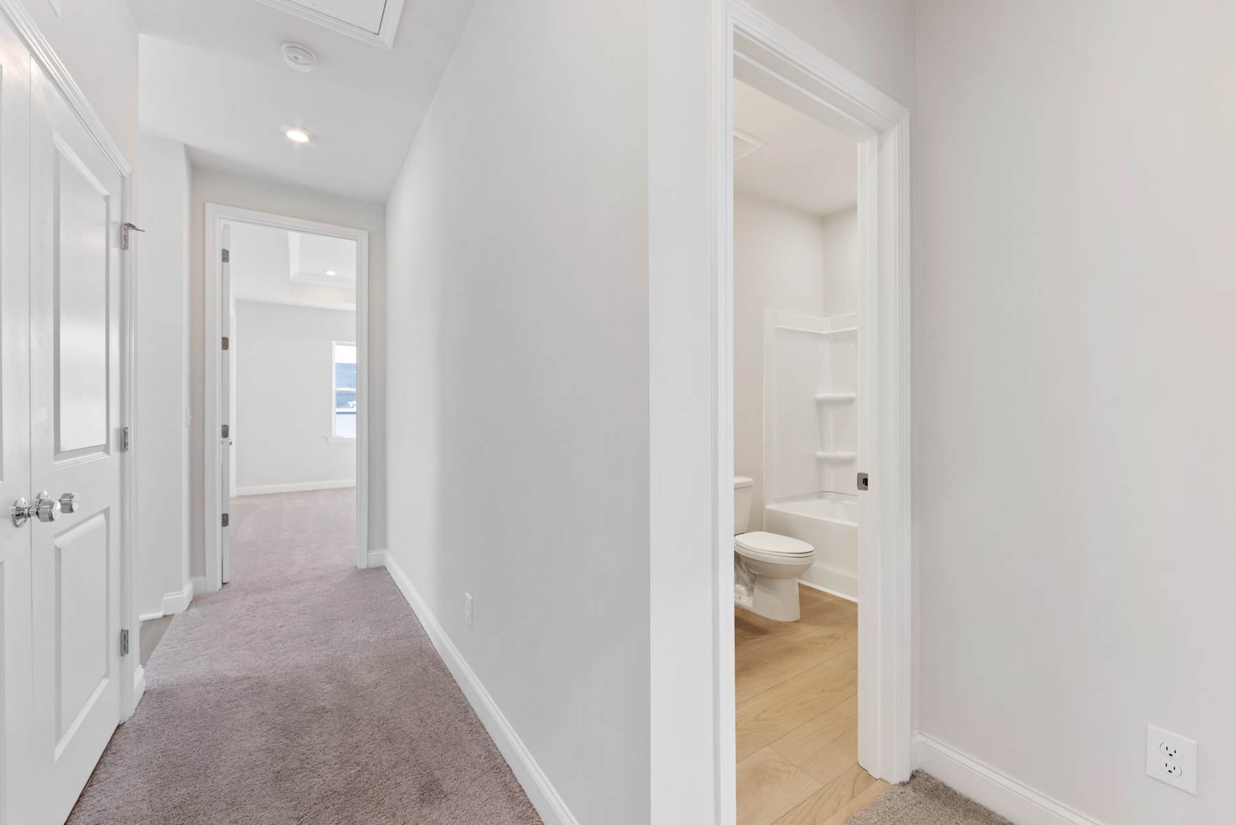 A bright, spacious hallway with white walls and a carpeted floor leads to an open doorway revealing a bathroom.