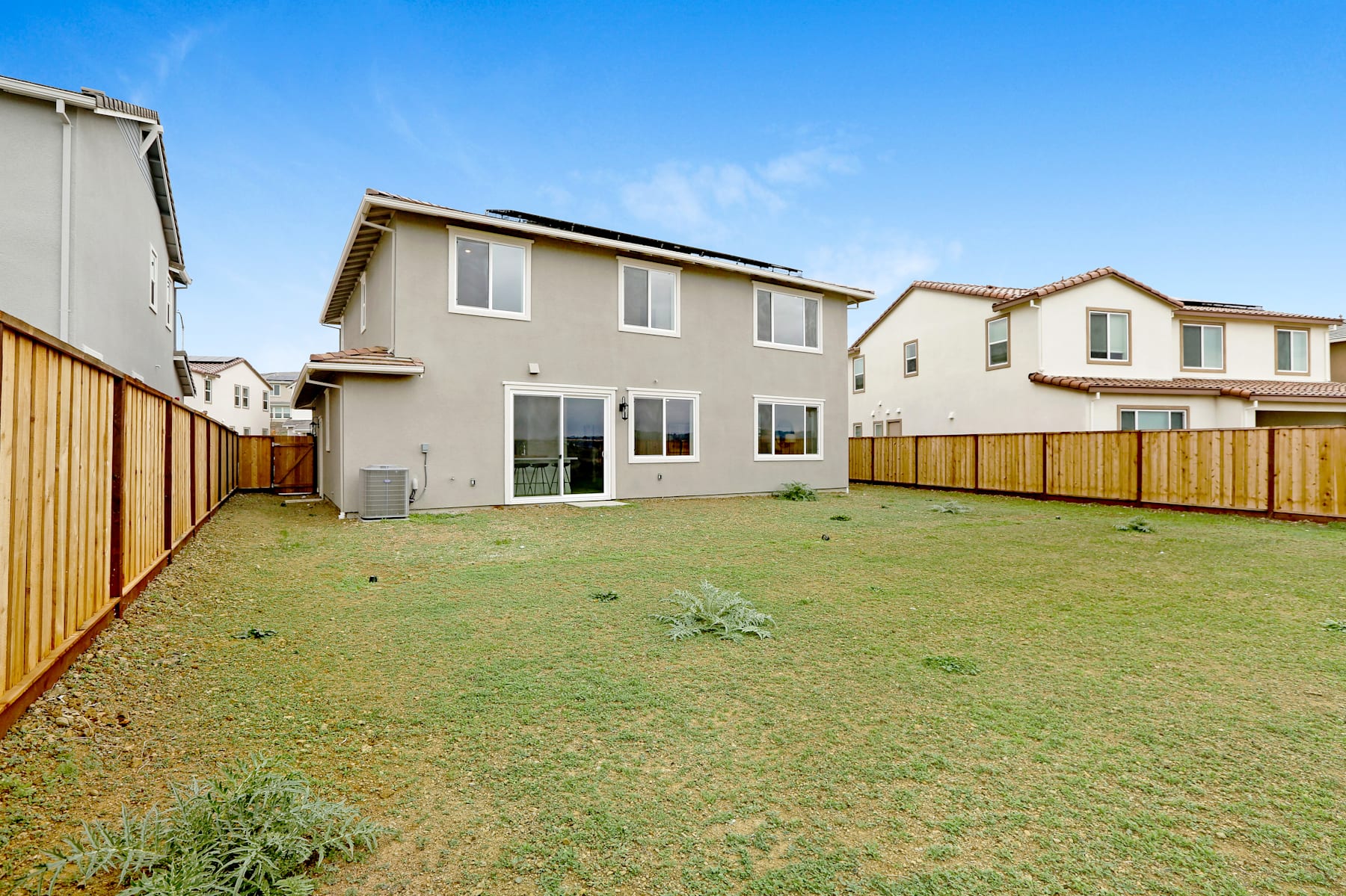 A residential neighborhood with newly constructed two-story houses, a grassy yard in the foreground, and a wooden fence separating the properties.