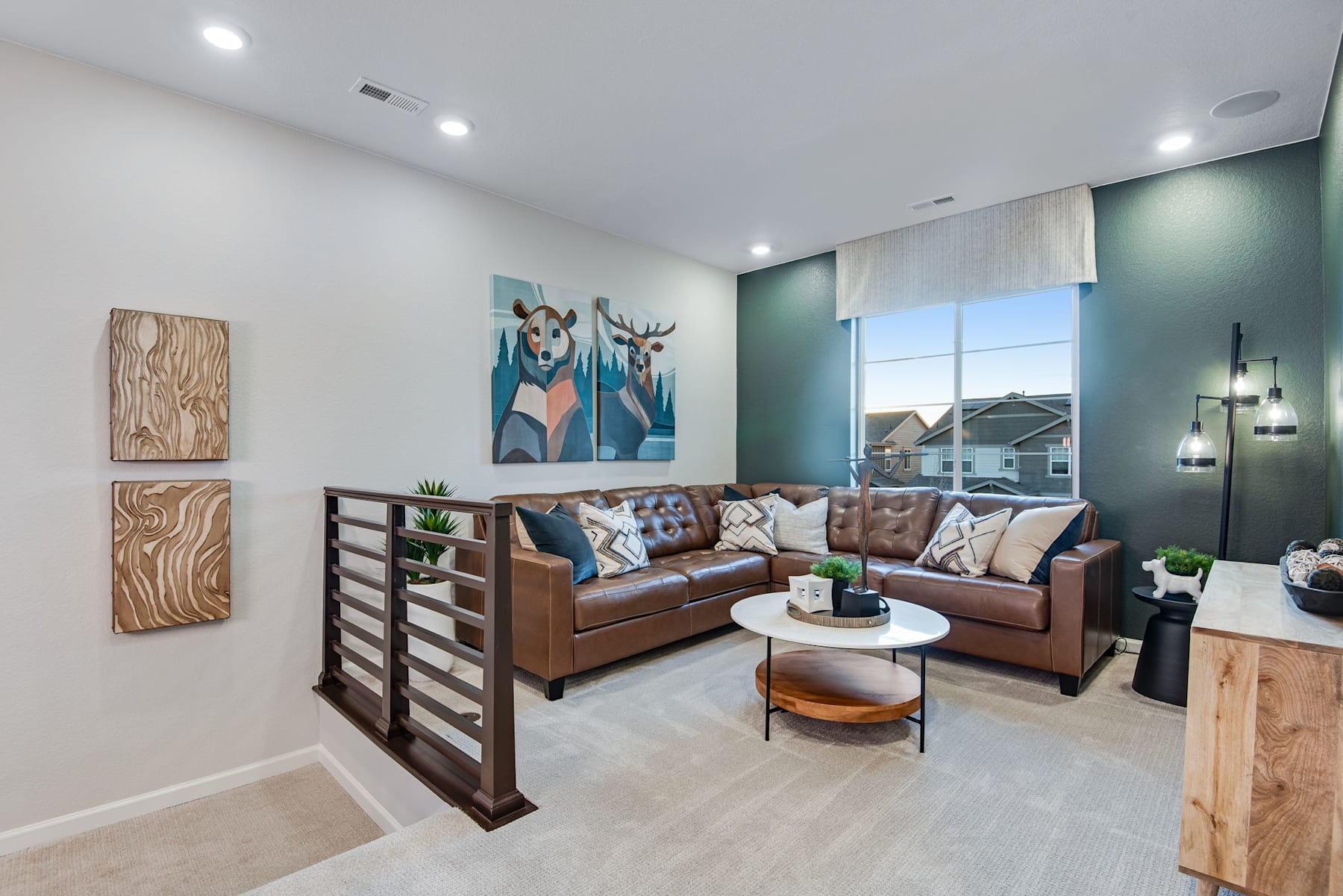 A cozy and modern living room with a large sectional sofa, wooden shelving, and artwork on the walls, set against a backdrop of large windows overlooking a residential neighborhood.