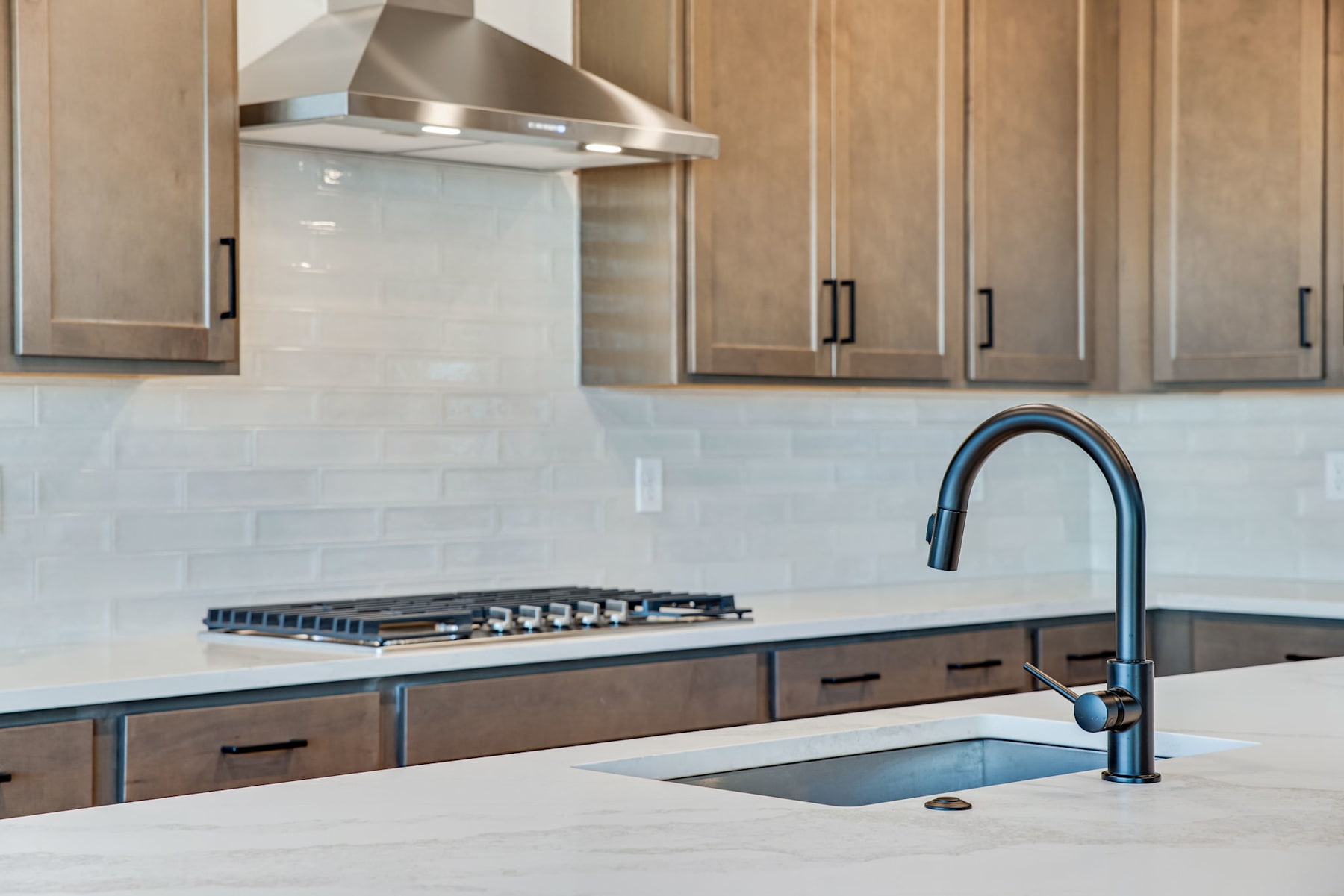 A modern kitchen with wooden cabinets, a stainless steel range hood, and a sink with a black faucet set against a tiled backsplash.