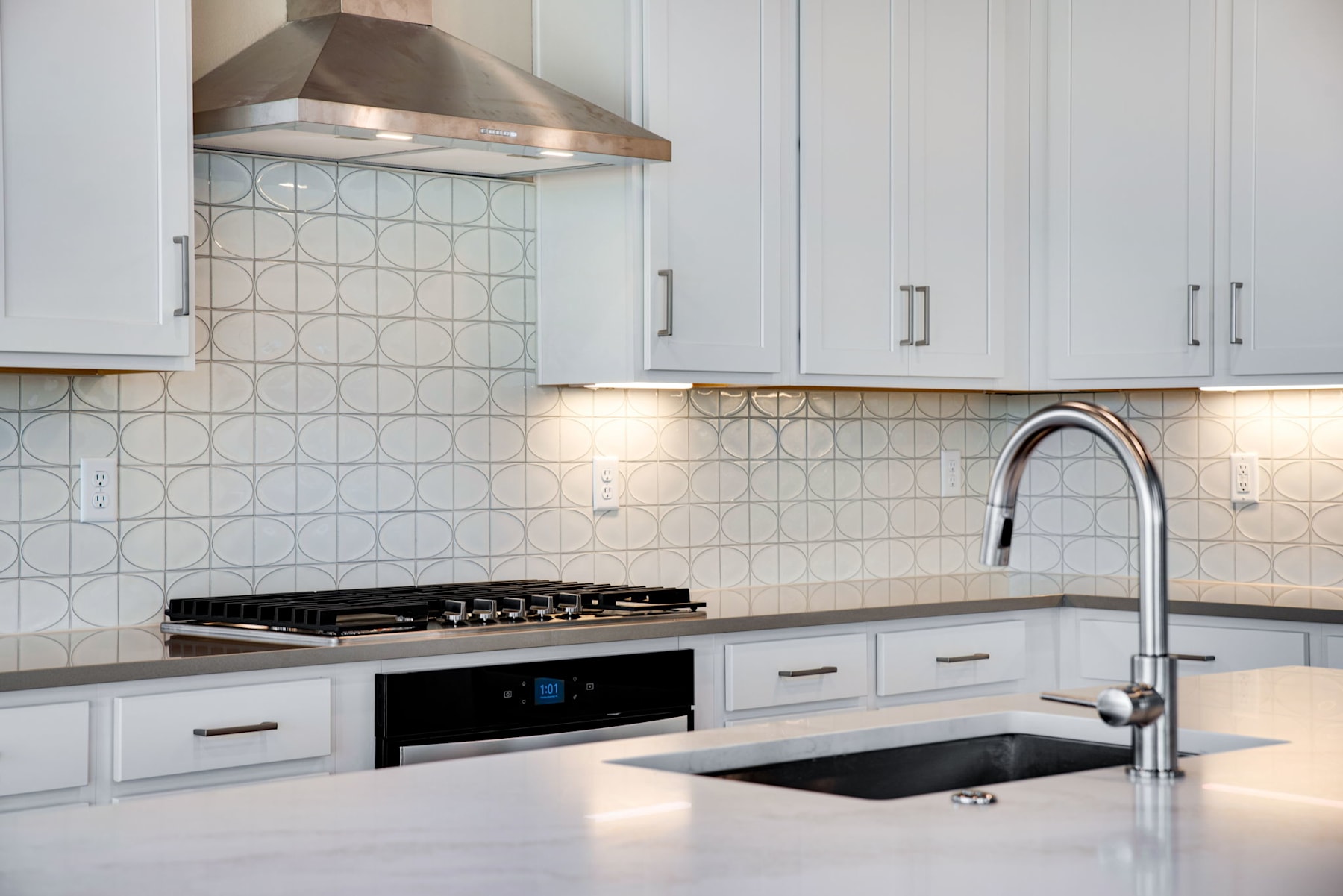 A modern, well-equipped kitchen with white cabinets, a stainless steel range hood, and a tiled backsplash, featuring a sink and faucet in the foreground.