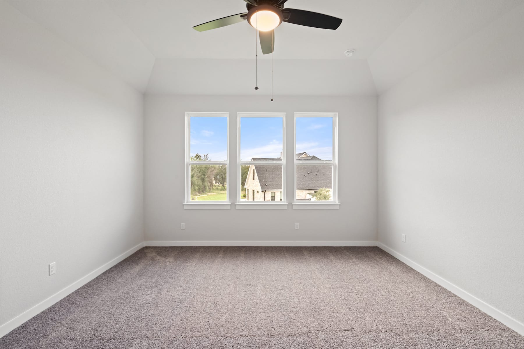 A sparsely furnished room with a ceiling fan, large windows overlooking a residential area, and a carpeted floor.