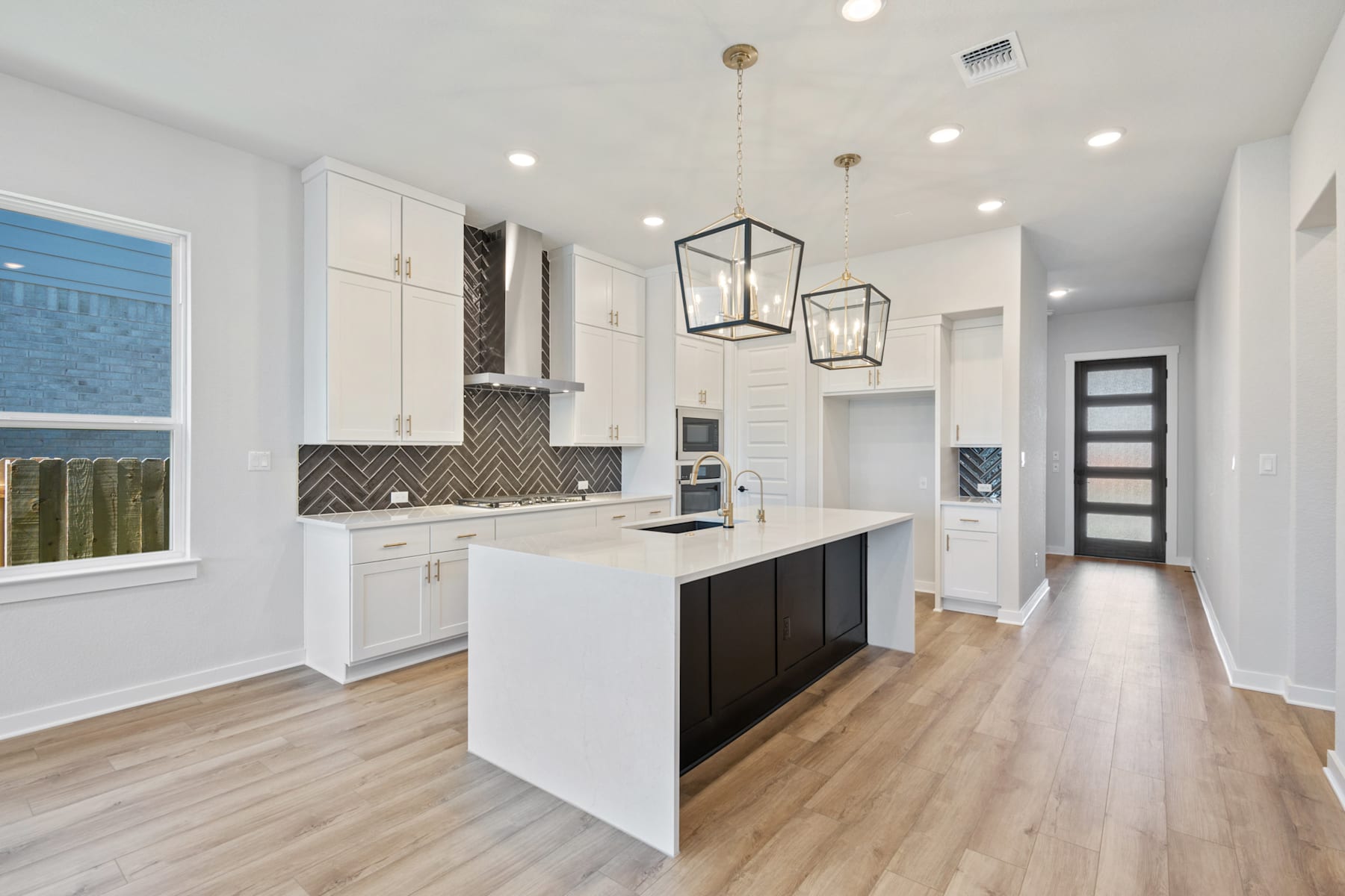 A modern, open-concept kitchen with white cabinets, a black island, and geometric pendant lights, leading into a hallway with a wooden floor.