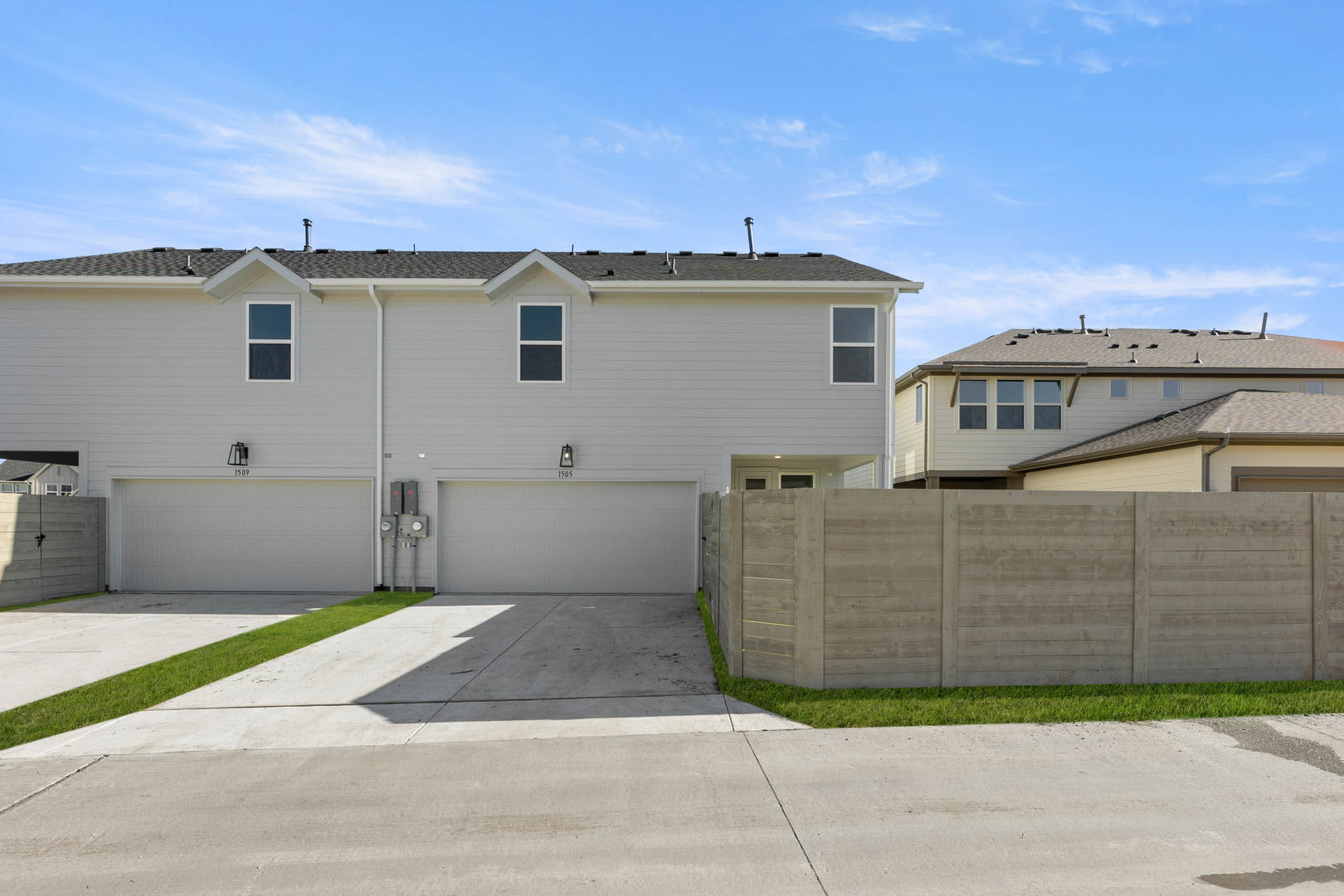 The image shows a residential neighborhood with a row of two-story townhouses, each with a garage and a small yard in the foreground. The sky is clear and blue, and there are some trees and other buildings visible in the background.