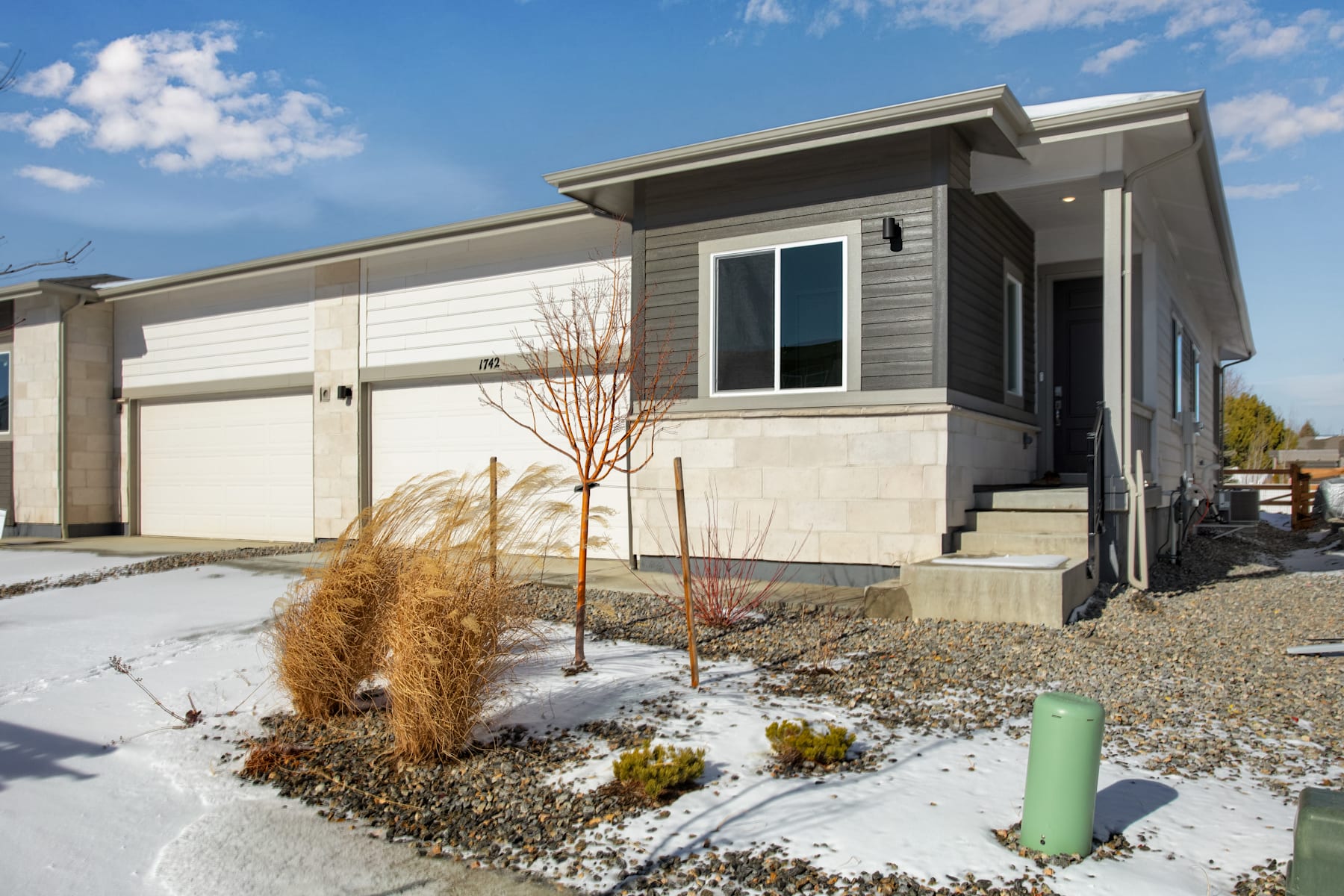 A modern, two-story residential home with a garage, surrounded by a landscaped yard with gravel, plants, and a small tree in the foreground.