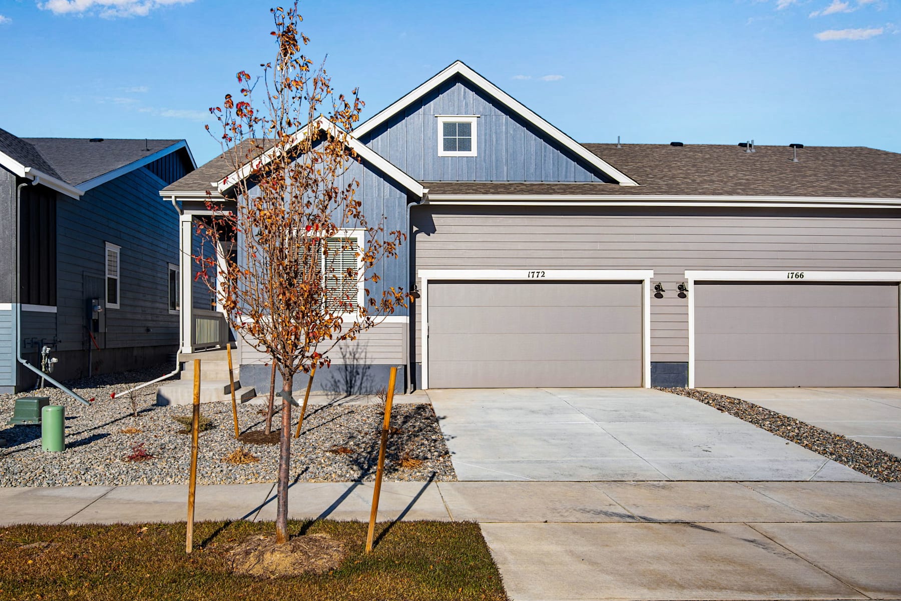 A modern two-story house with a garage, surrounded by a grassy lawn and a few young trees in the foreground, set against a clear blue sky with scattered clouds.