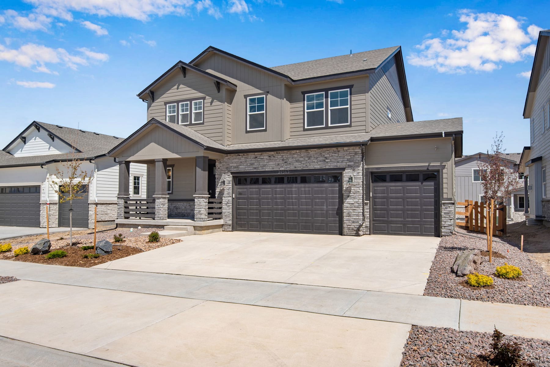 A two-story residential house with a gray exterior, a stone facade, and a double garage door sits in a well-landscaped yard with a paved driveway and a clear blue sky with scattered clouds in the background.