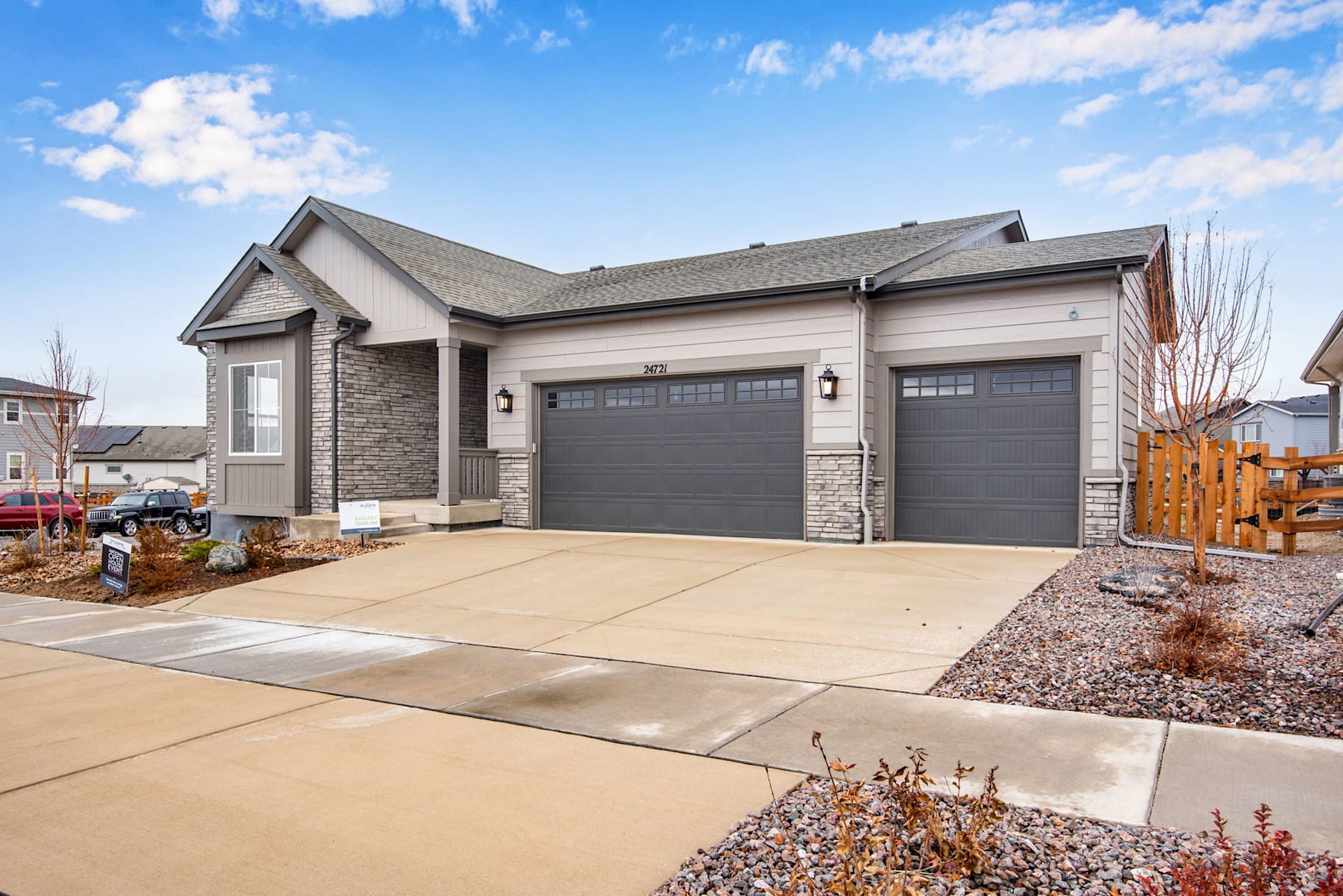 A two-story residential house with a gray exterior, a tiled roof, and a paved driveway leading to a pair of garage doors. The house is set against a backdrop of a clear blue sky with scattered clouds.
