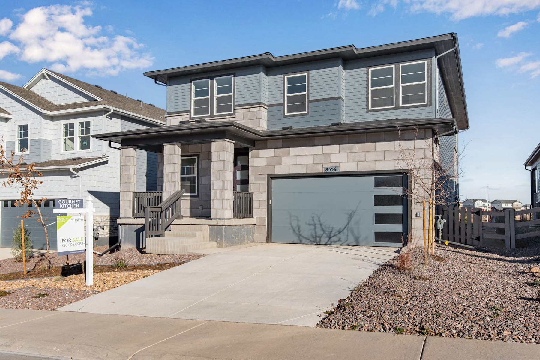 A modern two-story house with a garage, surrounded by a paved driveway and a grassy yard, set against a backdrop of cloudy blue sky.