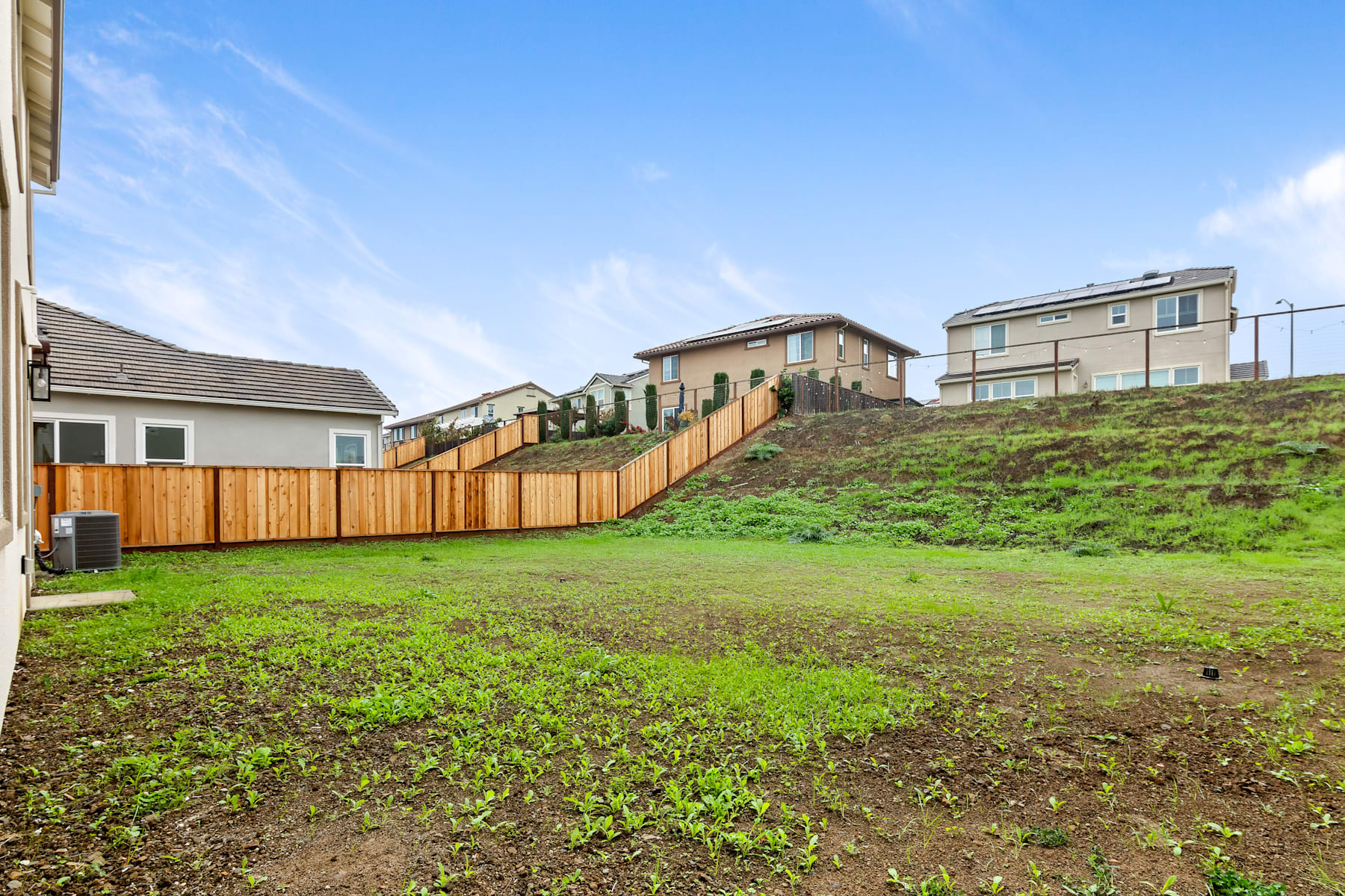 A grassy yard in the foreground with a wooden fence, leading up to a row of residential houses on a hillside in the background under a clear blue sky.