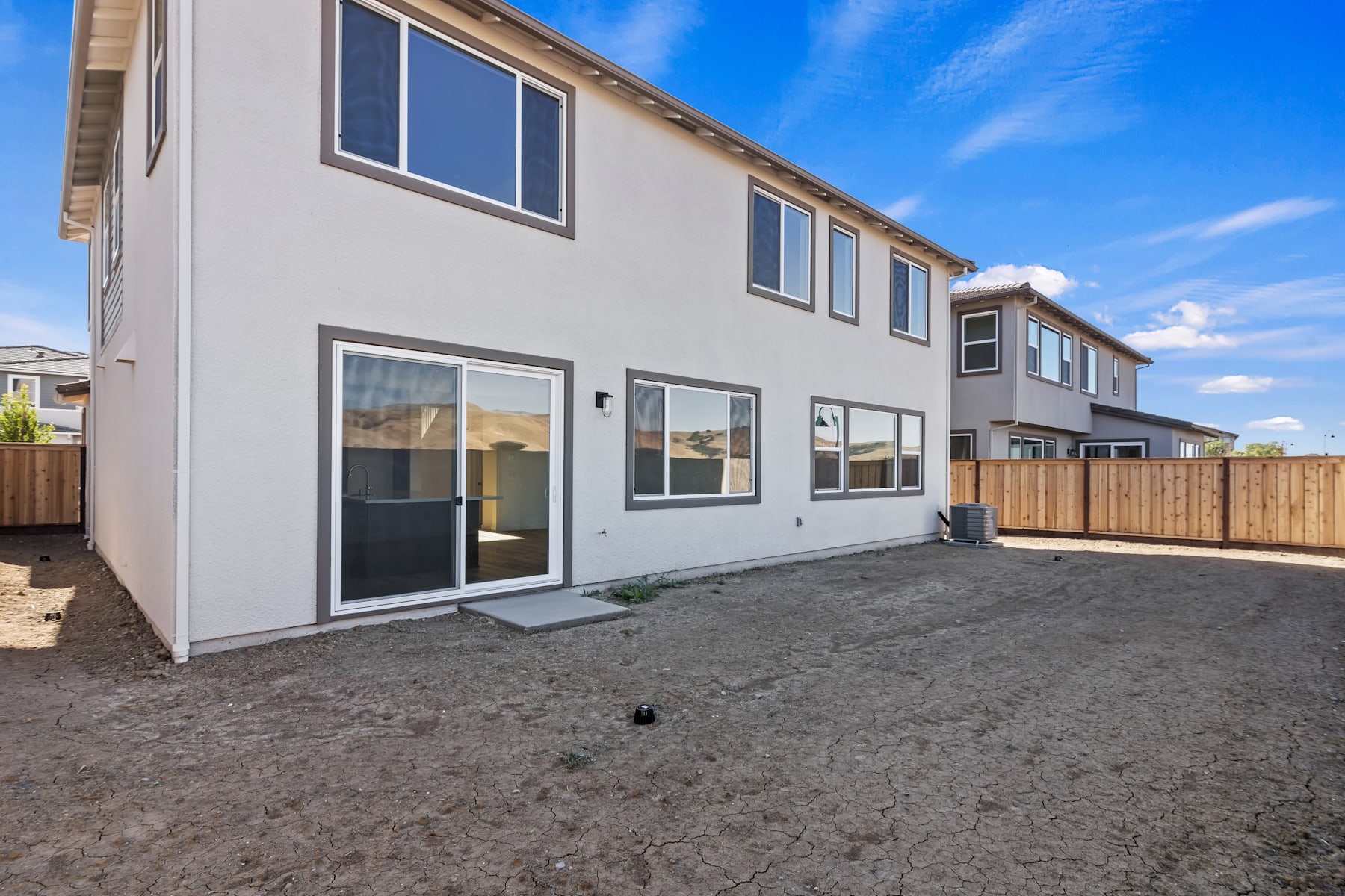 A newly constructed multi-unit residential building with a gray exterior, surrounded by a gravel driveway and a wooden fence in the foreground, set against a clear blue sky.