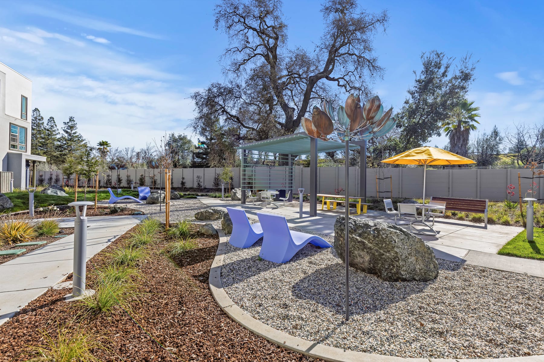 A modern outdoor seating area with colorful furniture, a large tree, and a whimsical metal sculpture in the foreground, surrounded by buildings and landscaping in the background.